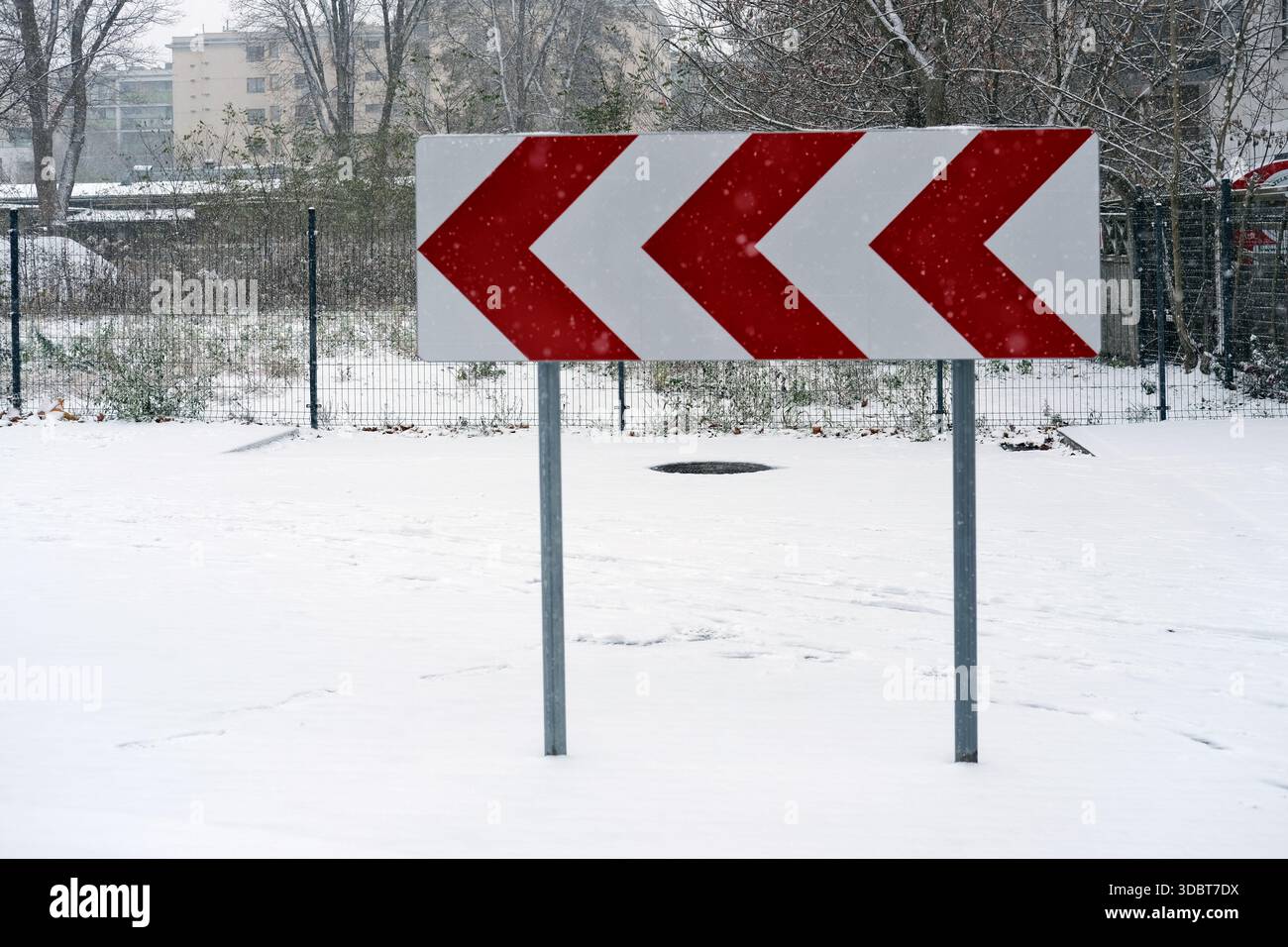 Richtungsschild mit roten und weißen Chevron-Pfeilen auf Metallpfosten, im Freien bei Schneefall fotografiert; schneebedeckter Boden. Stockfoto