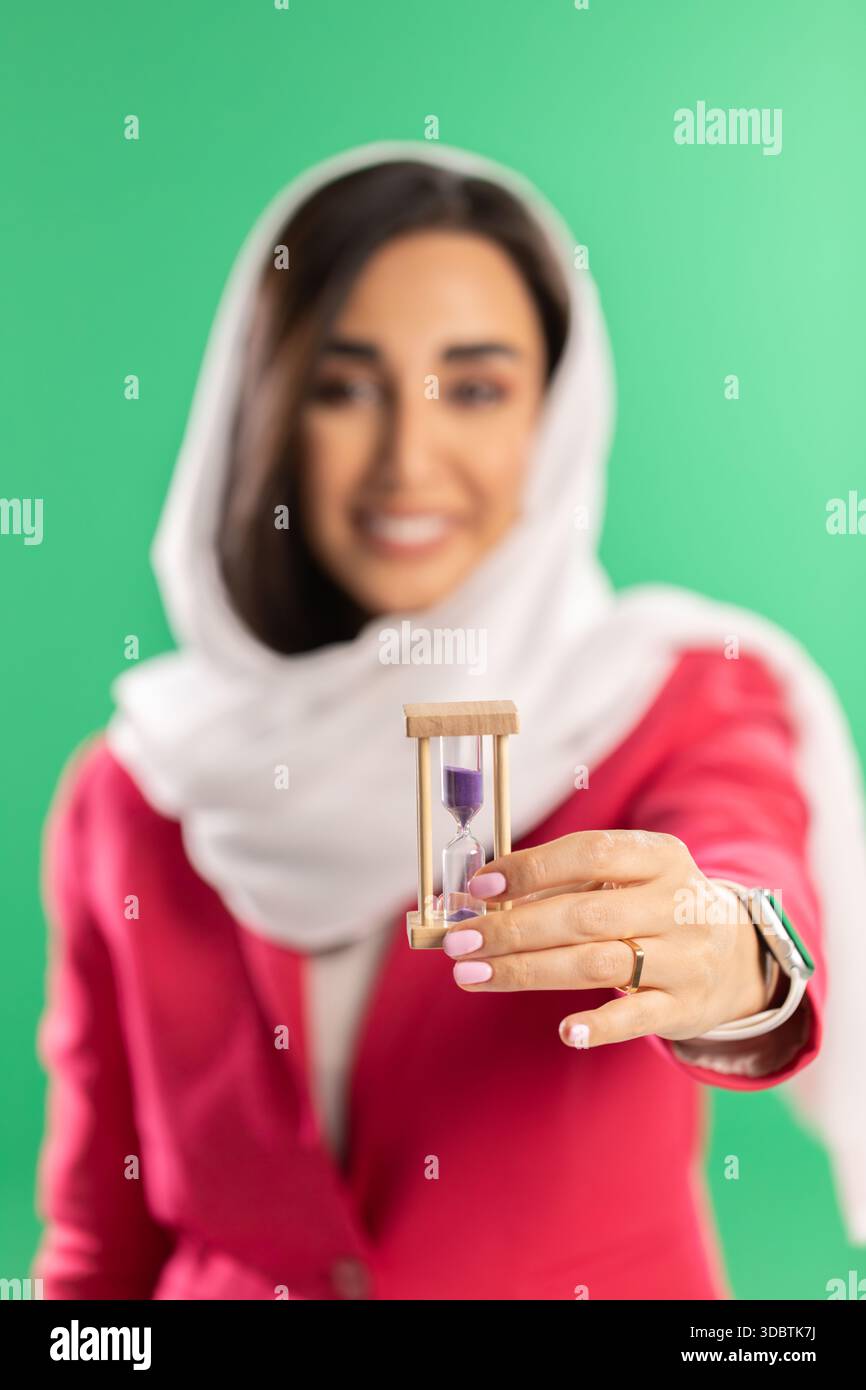 Eine Frau in weißem Kopftuch und rosafarbenem Blazer hält eine kleine hölzerne Sanduhr mit violettem Sand vor einem soliden grünen Hintergrund. Stockfoto