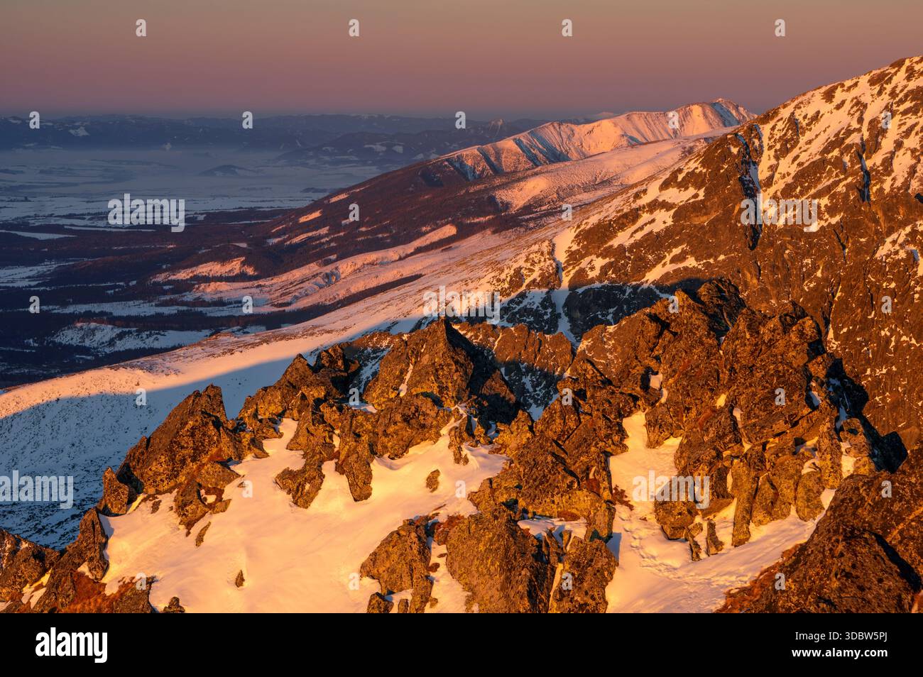 Der Blick auf zerklüftete, sonnenverwöhnte Gipfel erhebt sich über einem schneebedeckten Tal, wo Schatten sich verlängern und goldenes Licht auf dem zerklüfteten Gelände tanzt, Vysoke Tatry, Stockfoto