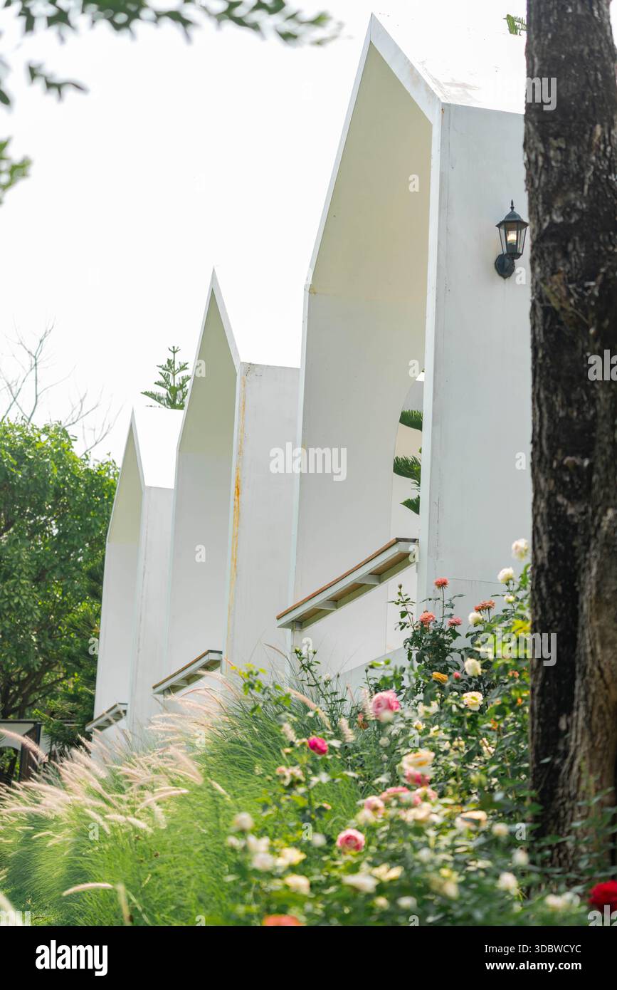 Flacher Blick auf moderne, weiße Giebeldächer. Umgeben von einem lebhaften Garten mit bunten Rosen und weichen Grasblumen unter einem hellen Stockfoto