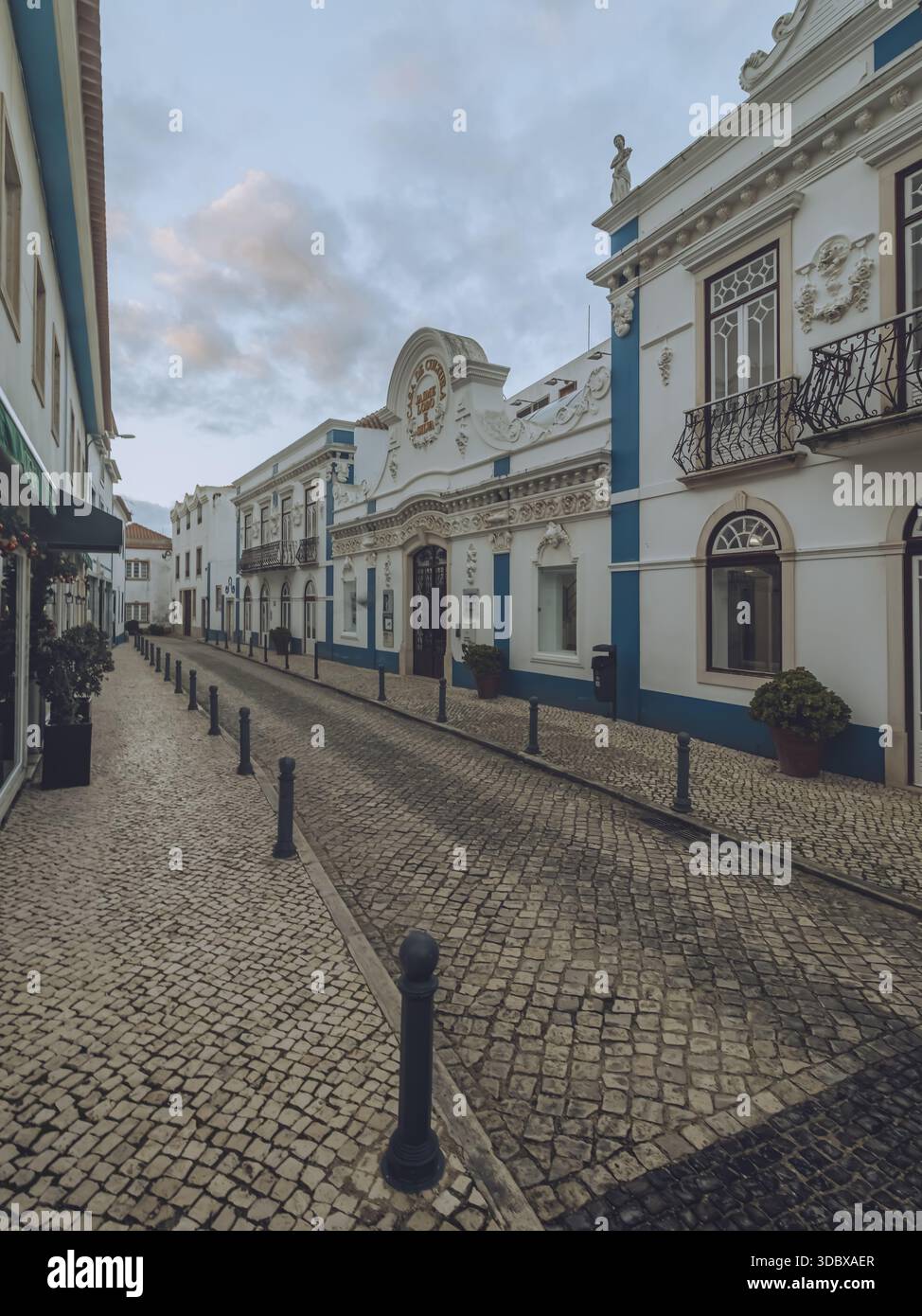 Blick auf die kopfsteingepflasterte Straße, die zu den alten weißen Gebäuden mit blauer Zierleiste unter einem bewölkten Himmel führt, Ericeira, Lissabon, Portugal. Stockfoto