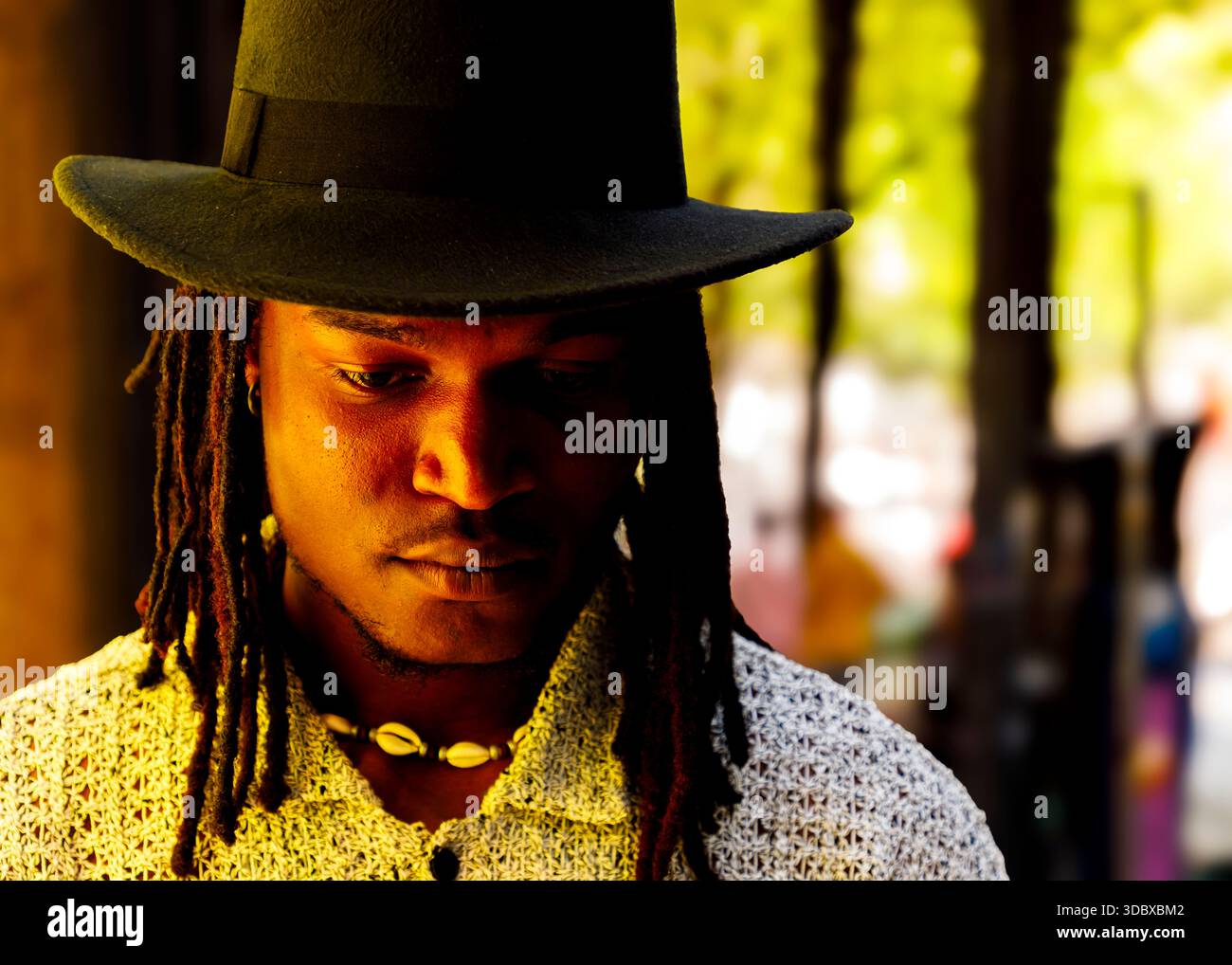 Ein Mann mit Dreadlocks, der einen schwarzen Hut und ein weißes Hemd trägt und nach unten blickt. Stockfoto