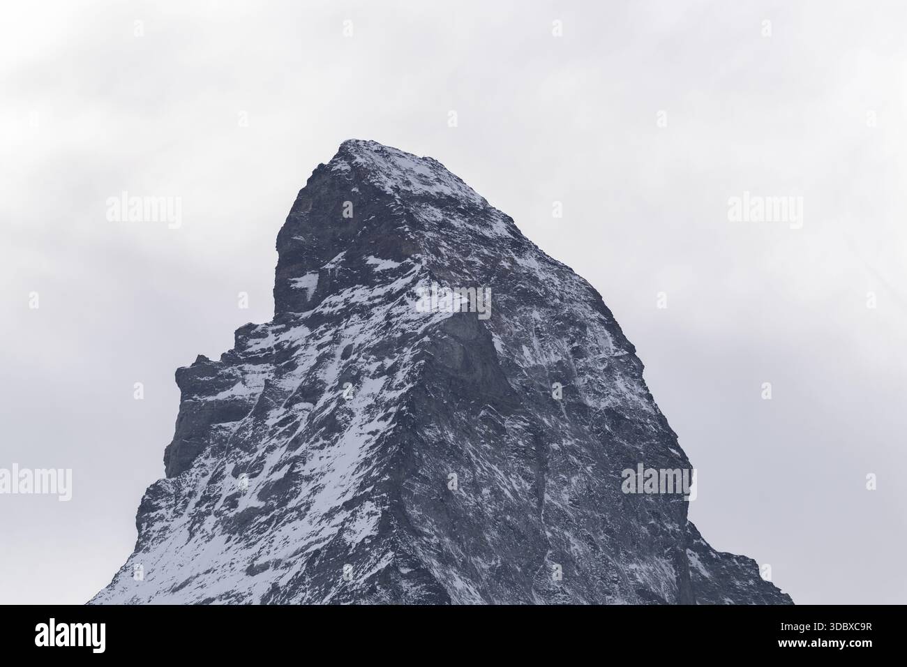 Blick auf einen schneebedeckten, zerklüfteten Berggipfel, der den hellen Himmel durchdringt, seine beeindruckende Präsenz bildet einen starken Kontrast zum weichen, diffusen Licht, Zermatt, Vala Stockfoto