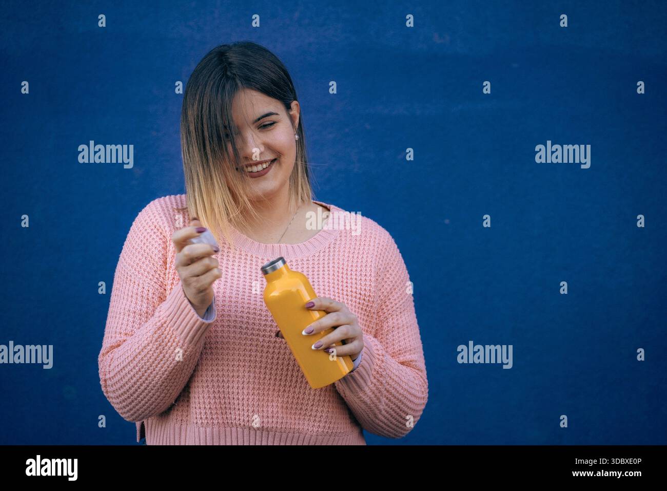 Lächelnde junge Frau in rosa Pullover, die eine gelbe wiederverwendbare Wasserflasche öffnet und vor einer leuchtend blauen Wand steht. Stockfoto