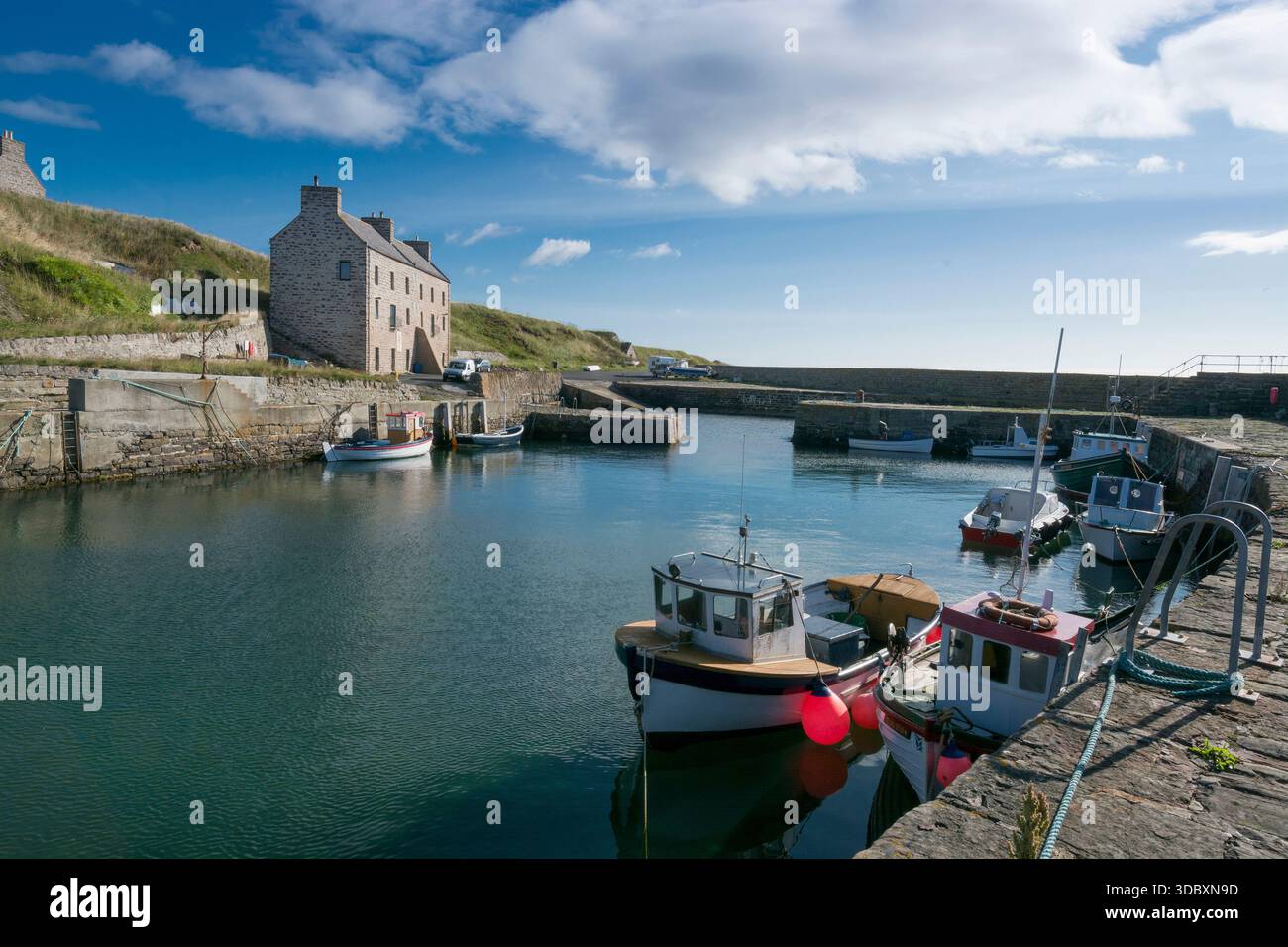 Eine ruhige Hafenszene von Caithness in Sinclair Bay mit Booten, die entlang eines Steinkaies unter blauem Himmel vor Anker liegen. Stockfoto