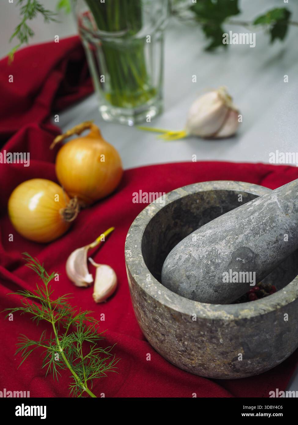 Steinmörtel und Pistill mit Pfefferhörnern, Knoblauch und Zwiebeln auf rotem textilem Hintergrund Stockfoto