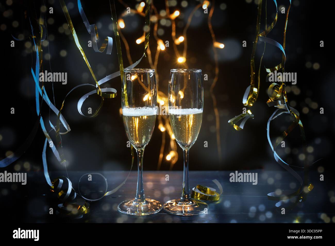 Zwei Sektflöten, goldene blaue Luftschlangen und Feenlichter vor dunklem Hintergrund mit Bokeh-Blasen, die eine Feiertagsparty wie nachts feiern Stockfoto