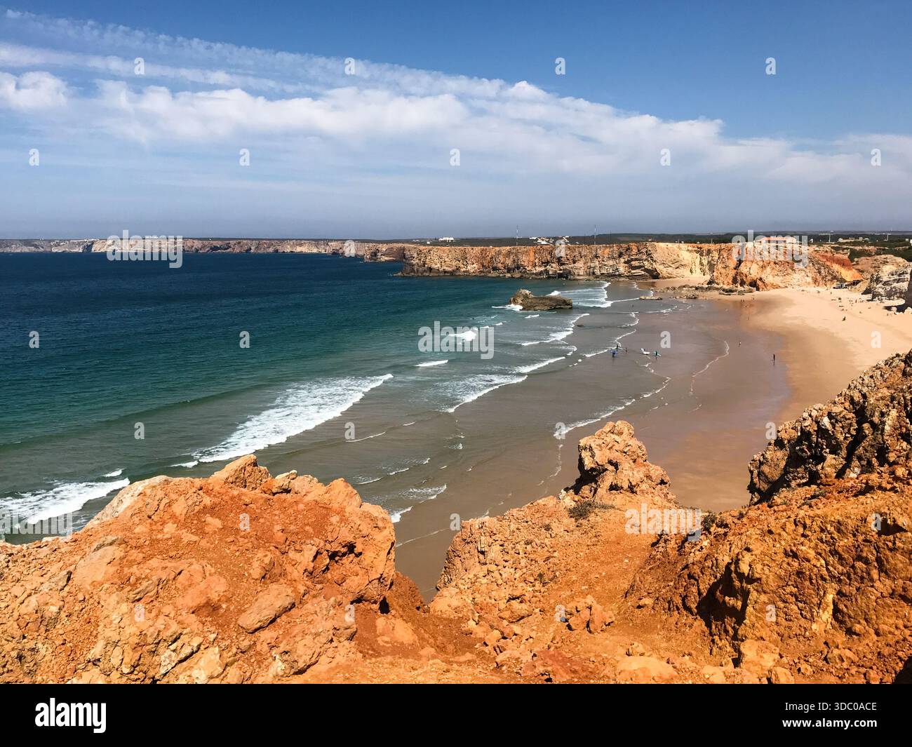 Eine Küstenlandschaft im Süden Portugals zeigt einen breiten Sandstrand, türkisfarbene Atlantikwellen und zerklüftete orangene Klippen unter einem klaren blauen Himmel. Stockfoto
