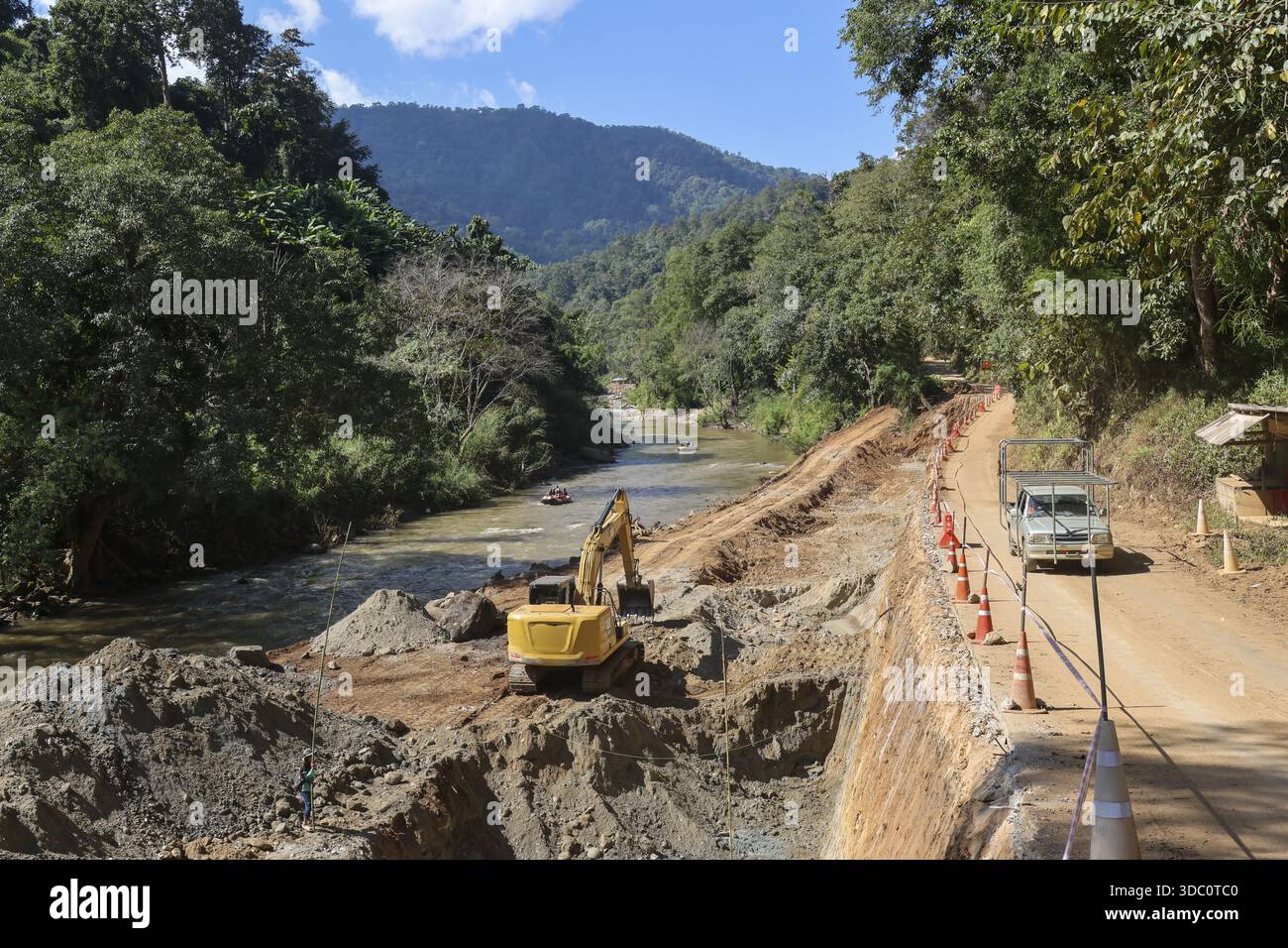 Schwerer gelber Bagger, der in der Nähe von Flussbaustellen Boden gräbt, mit grünem Berghintergrund, der den industriellen Fortschritt und massive Erdarbeiten zeigt Stockfoto