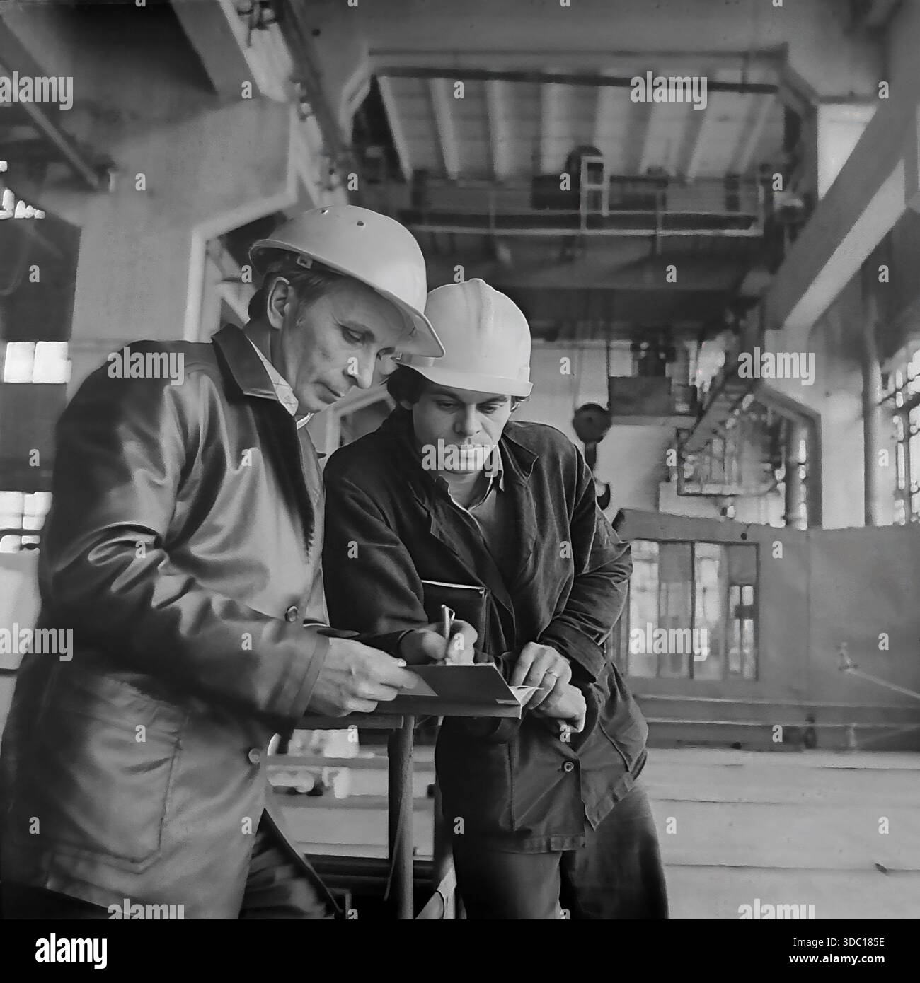 Sloviansk, Ukrainische SSR, UdSSR, 1985. Ein Archivfoto aus dem AIZ-Werk (Armaturno-Izolyatorny Zavod). Zwei Mitarbeiter von Bauhelmen, wahrscheinlich auch Agatuev, besprechen Produktionsaufgaben in der Werkstatt. Der erfahrene Spezialist in einer Lederjacke checkt Notizen in einem Notizbuch seines jüngeren Kollegen. Der Hintergrund zeigt die Größe der Industrieanlage mit Kränen und Anlagen. Eine lebhafte Episode des Arbeitsprozesses im friedlichen Donbass während der Sowjetzeit. Stockfoto