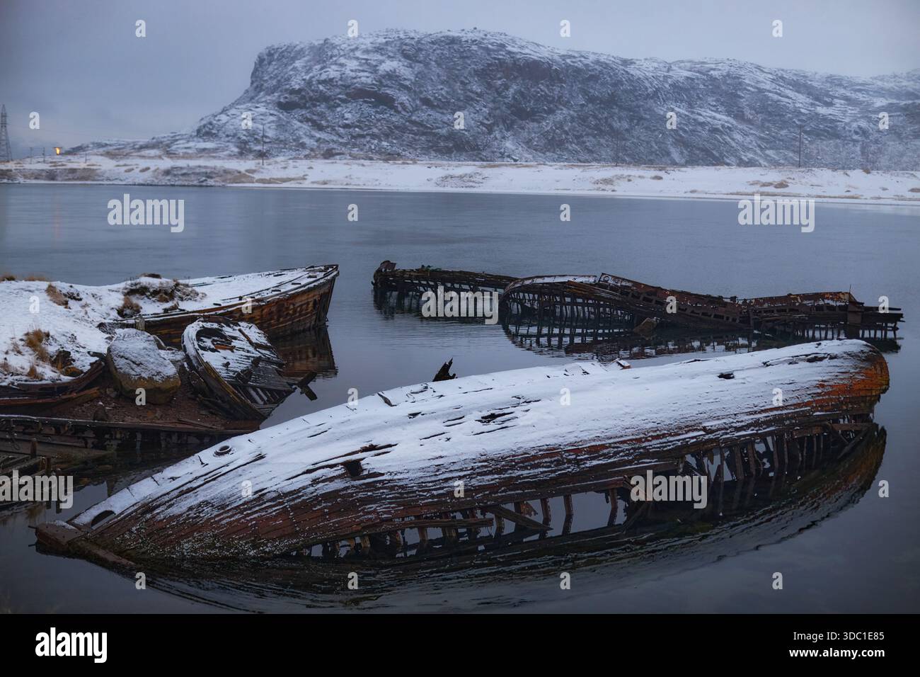 Ruinierte Boote ruhen in Teriberka, Russland, halb untergetaucht im Wasser mit Schnee auf ihnen und Bergen im Hintergrund während der frühen Morgenstunden. Stockfoto