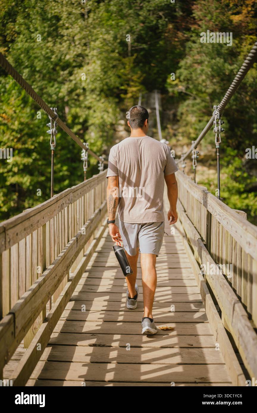 Rückblick eines jungen Mannes, der über eine Hängebrücke läuft und Wasser hält Stockfoto