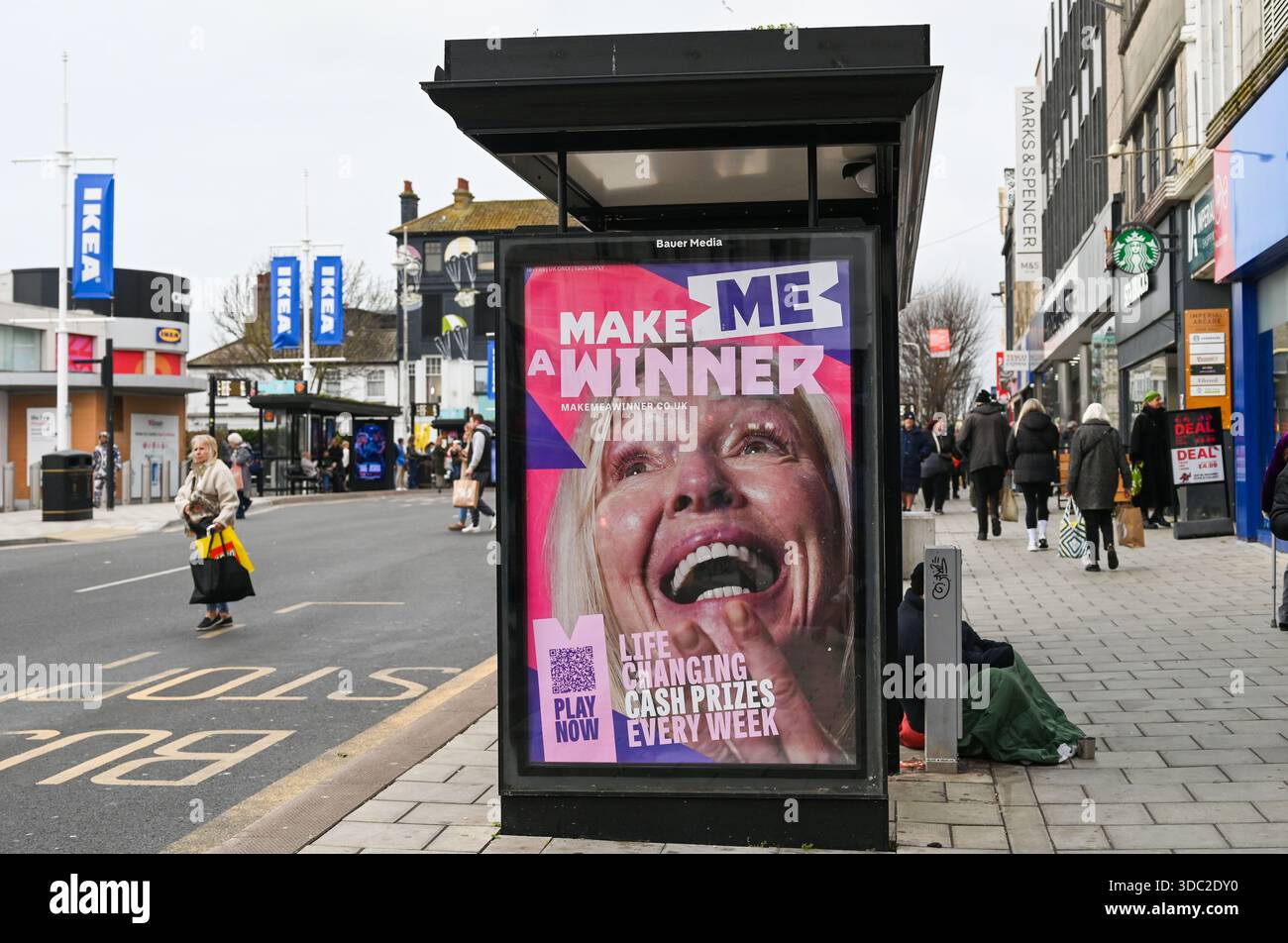 Eine elektronische Werbetafel von Bauer Media, die die National Lottery in einer Bushaltestelle in der Western Road Brighton zeigt, mit einem Mann, der außerhalb des Dezember 2025 um Geld bittet Stockfoto