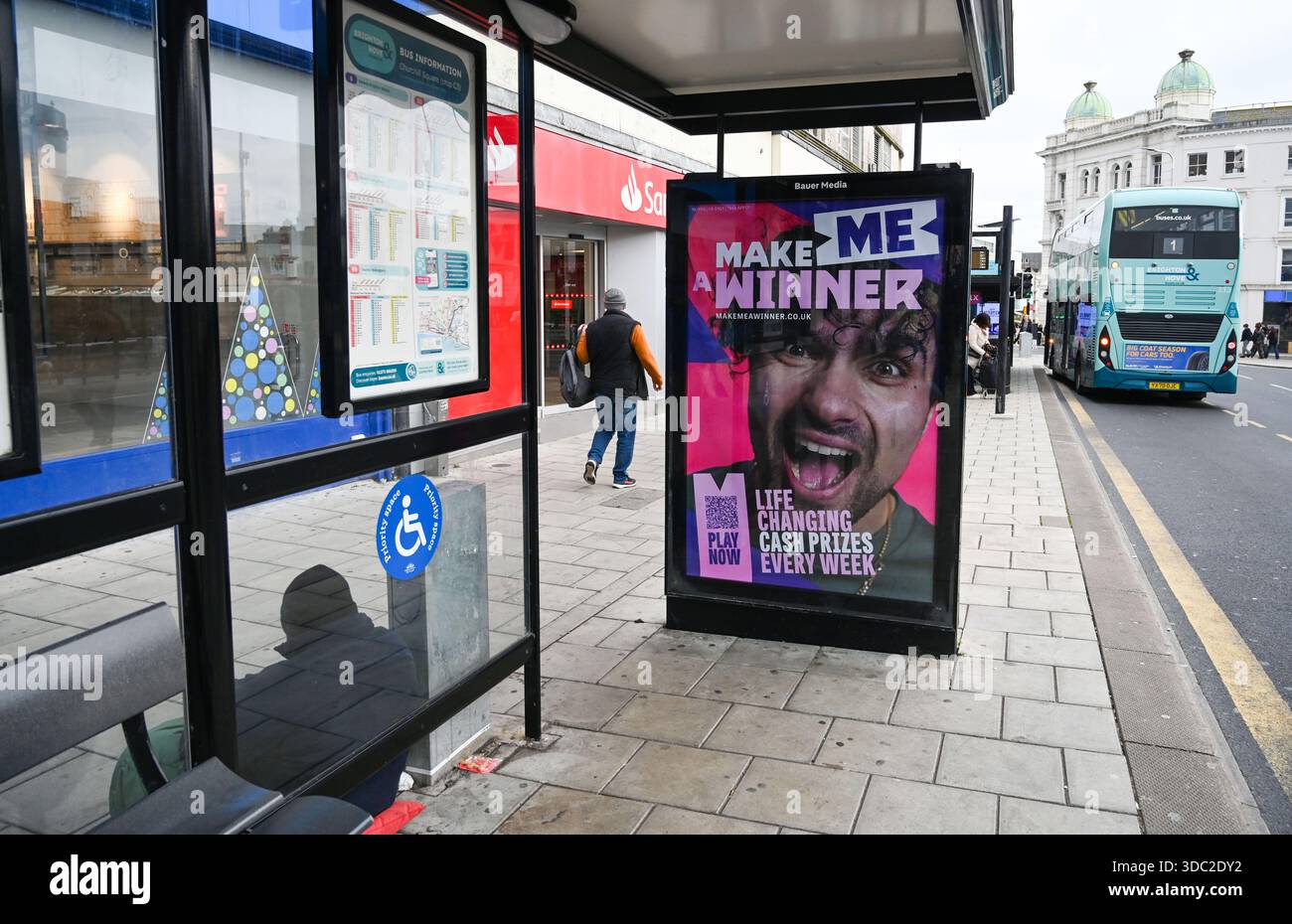 Eine elektronische Werbetafel von Bauer Media, die die National Lottery in einer Bushaltestelle in der Western Road Brighton zeigt, mit einem Mann, der außerhalb des Dezember 2025 um Geld bittet Stockfoto