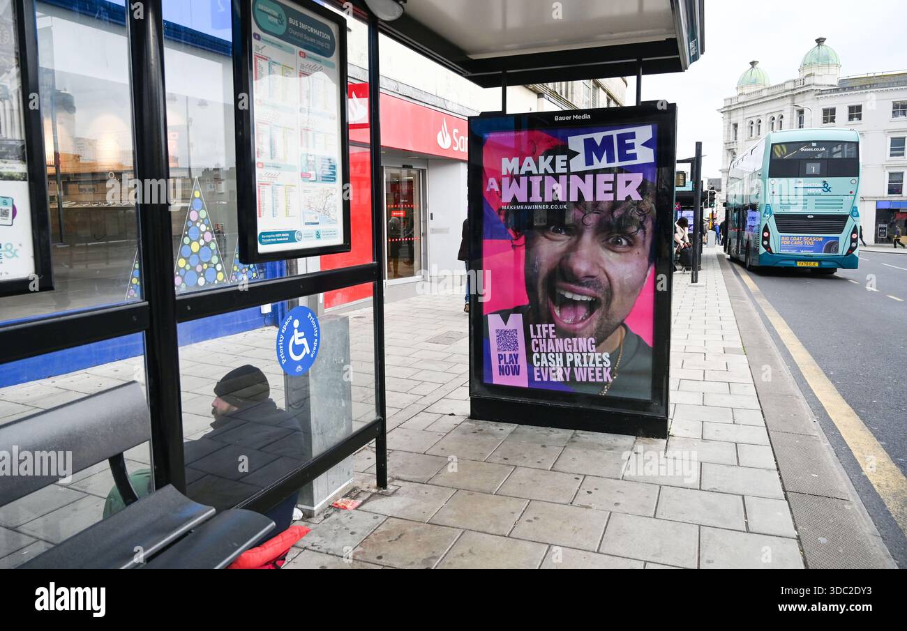 Eine elektronische Werbetafel von Bauer Media, die die National Lottery in einer Bushaltestelle in der Western Road Brighton zeigt, mit einem Mann, der außerhalb des Dezember 2025 um Geld bittet Stockfoto
