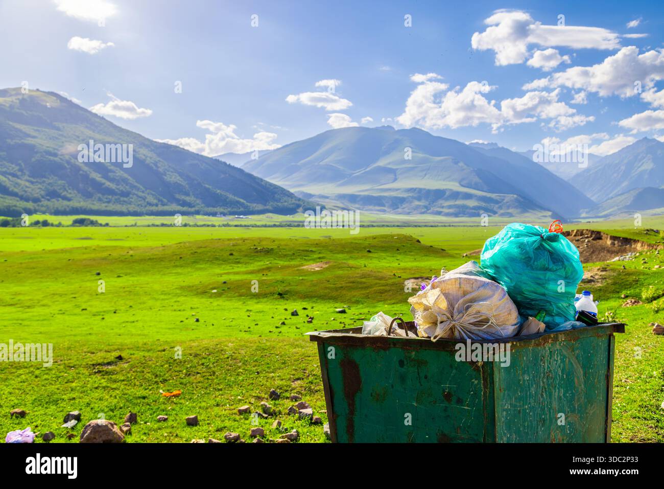 Grüner Abfallbehälter aus Metall überläuft mit hellbraunem Netz und türkisfarbenen Plastiktüten. Die landschaftlich reizvolle Lage im Bergtal unterstreicht die Umweltbelange. Stockfoto