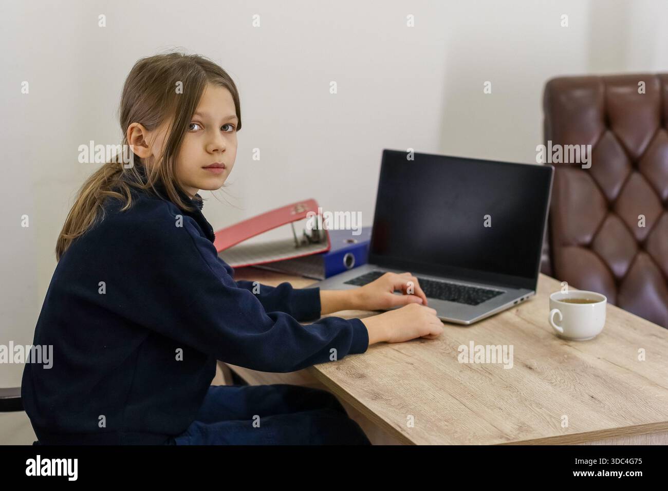 Eine junge Frau dreht den Kopf leicht um und sitzt am Tisch mit Händen in der Nähe des Laptops, farbenfrohen Mappen und Getränken in neutraler Innenumgebung. Stockfoto