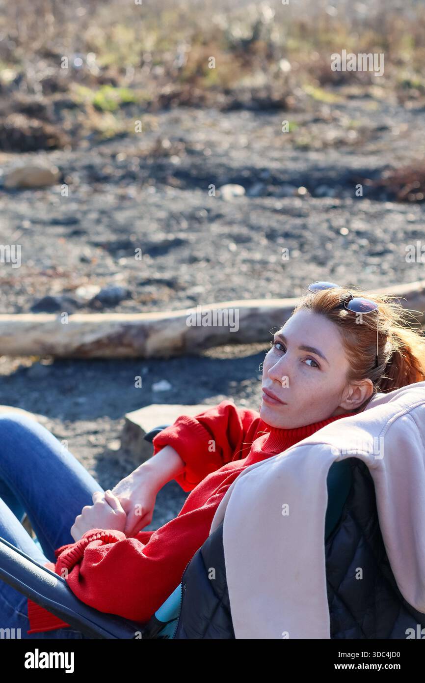 Vertikales Porträt einer rothaarigen Frau im Rollkragenpullover, liegend im Klappstuhl auf felsigem Boden, seitlich mit nachdenklichem Ausdruck. Stockfoto
