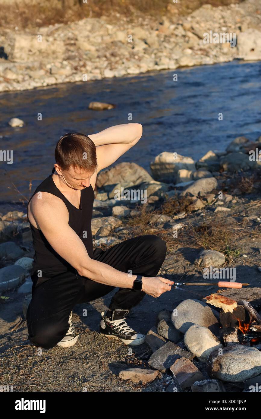 Nahaufnahme eines Mannes in sportlicher Kleidung, der sich ausdehnt, während er Wurst auf Metallspießen über glühender Lagerfeuerheuere auf Kies am Fluss kocht, was an Outdoor r erinnert Stockfoto