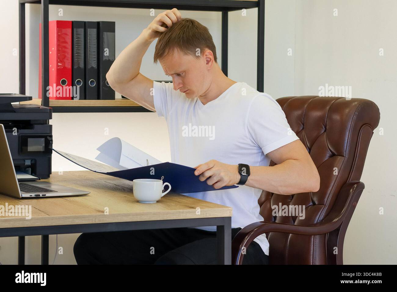 Verwirrter Arbeiter Hand am Haar, während er blaue Ordner am Arbeitsplatz mit Technik und Getränk untersucht Stockfoto