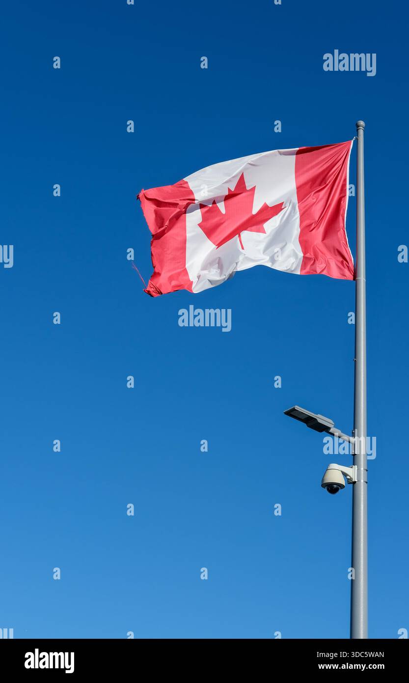 Flacher Außenanblick auf die rot-weiße Kanadas-Flagge auf dem Metallmast, sichtbare Stoffstruktur, montierte Straßenlaterne und Dome-Sicherheitskamera. Stockfoto