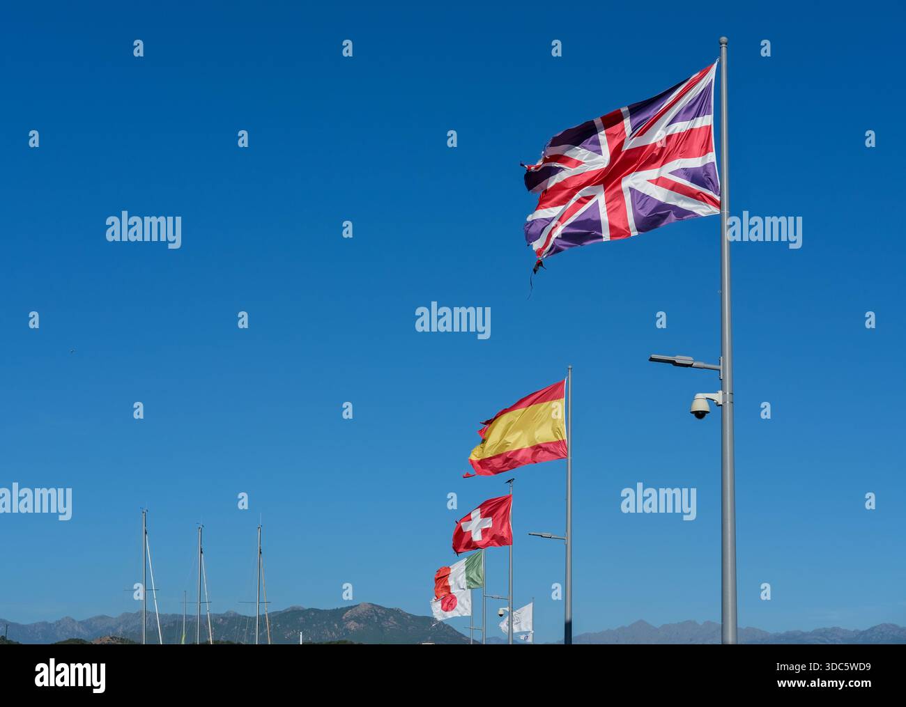 Flaggen aus Großbritannien, Spanien, der Schweiz, Italien und Japan auf hohen Metallpfählen montiert, aus niedrigem Winkel aufgenommen, klarer blauer Himmel, starkes Tageslicht. Stockfoto