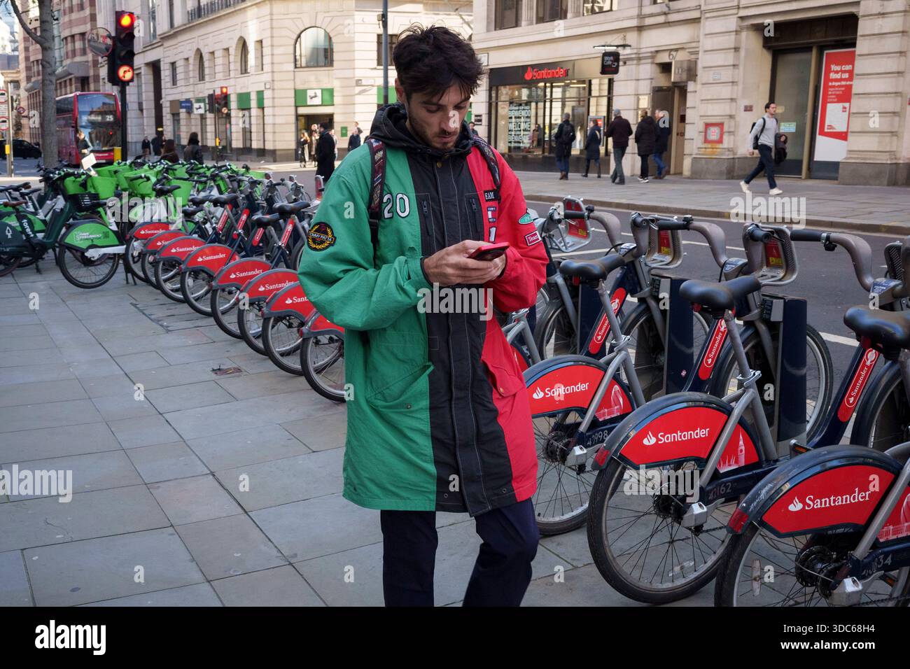 Ein Mann in einem grünen und roten Mantel läuft am 19. Dezember 2025 in London, dem Finanzviertel der Hauptstadt, an Lime und Santander Leihfahrrädern vorbei. Stockfoto
