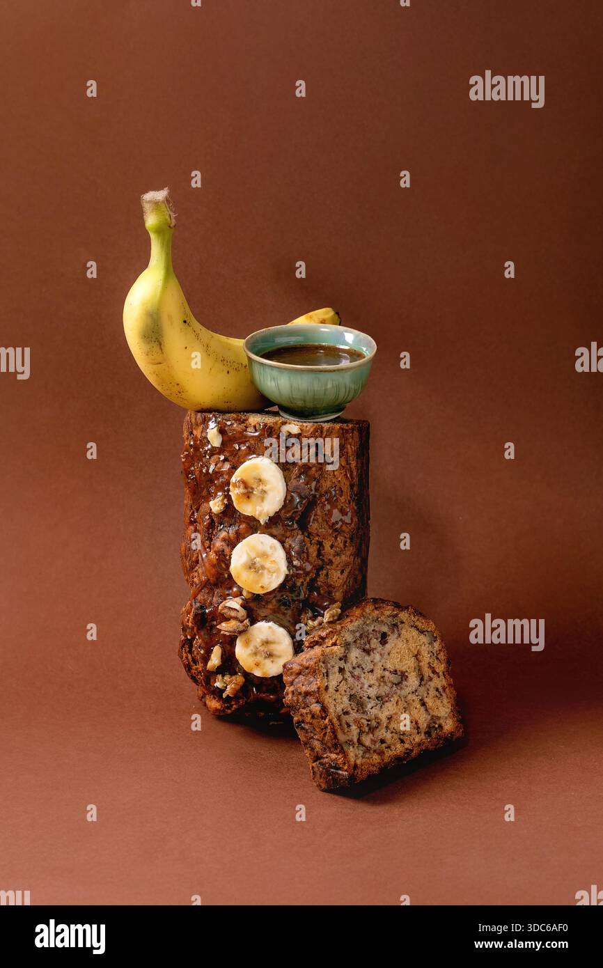 Modernes Stillleben mit hausgemachtem Bananenbrot. Brot mit Walnüssen, bestäubtem Karamell und frischer Banane auf braunem Hintergrund. Banane und Soße Stockfoto