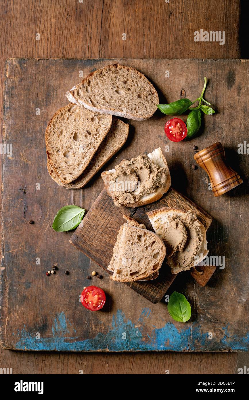 Hausgemachte Brotscheiben mit Leberpastete auf rustikalem Holzbrett. Frische Basilikumblätter, Kirschtomaten und Pfeffermühle. Traditionelle Bio-Frühstückszutaten Stockfoto