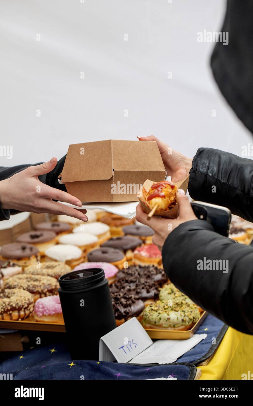 Person, die bunte Donuts und Gebäck am Street Food Stand kauft. Hände teilen sich Bastelbox mit süßen Leckereien. Verschiedene glasierte Donuts auf dem Präsentationsfach. C Stockfoto