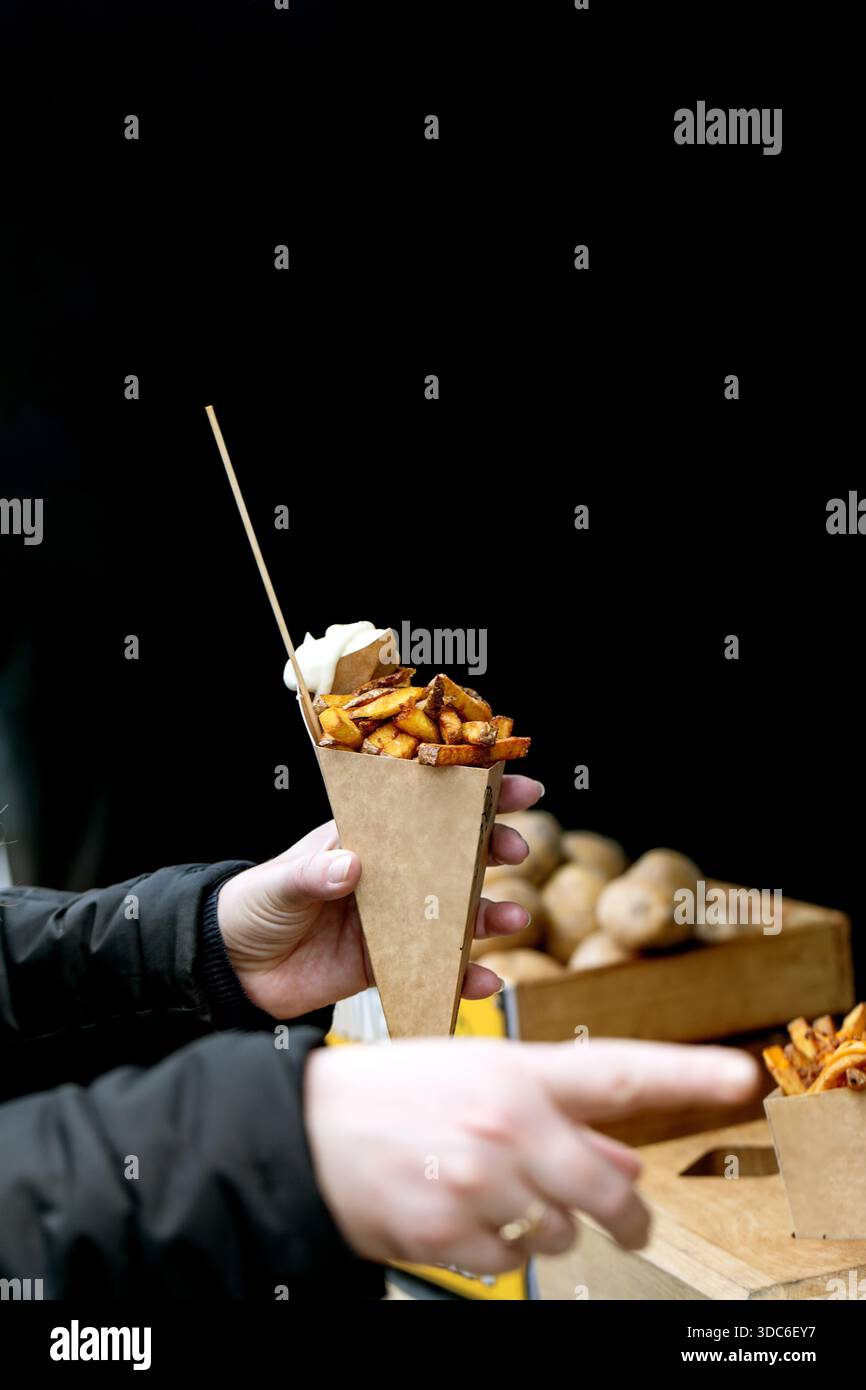 Hand hält Papierkegel mit gebratenen Kartoffeln und Soße auf dem Street Food Market. Street-Food-Take-away-Konzept vor schwarzem Hintergrund mit Food-Truck-Männern Stockfoto