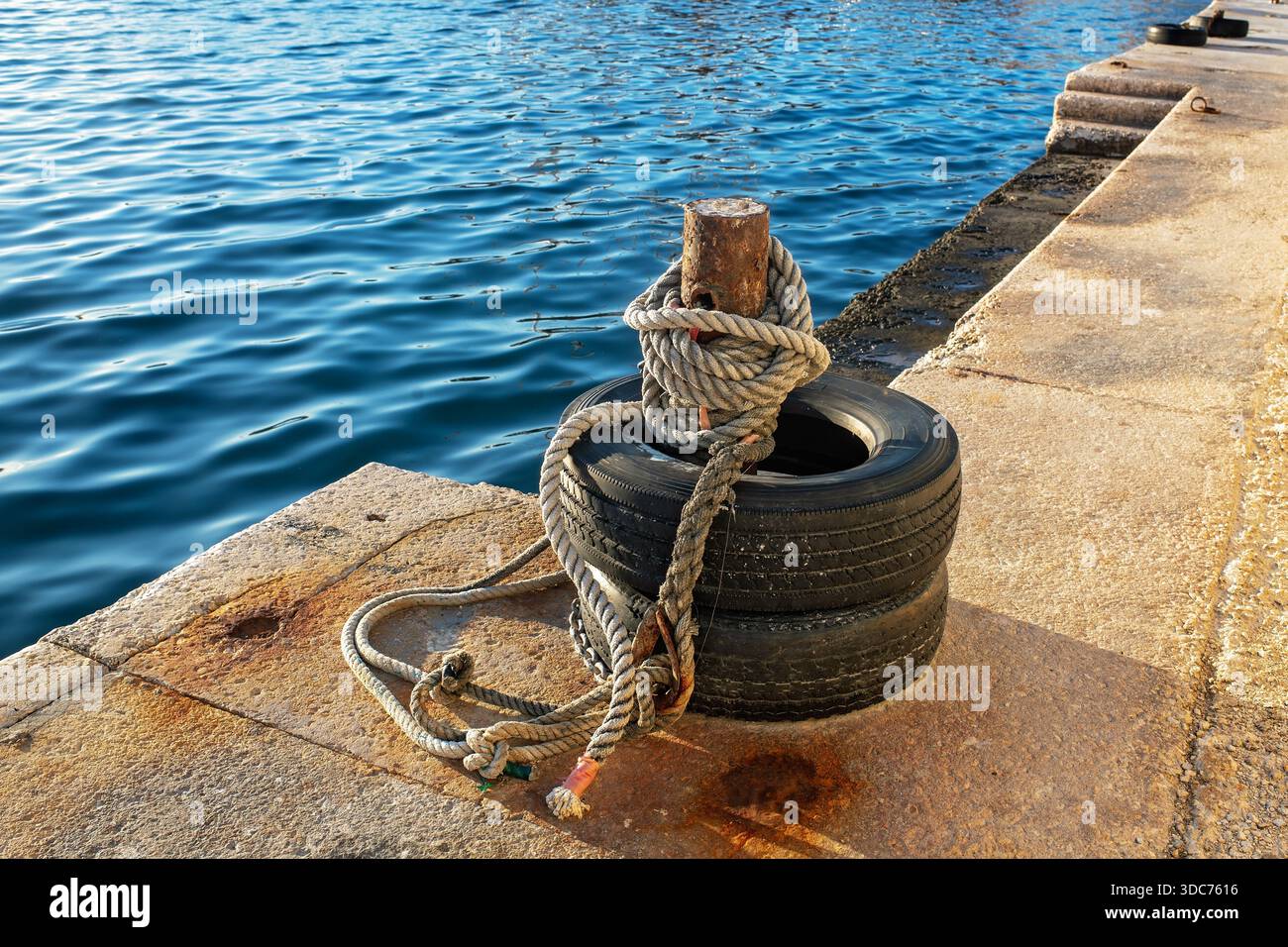 Nahaufnahme eines rostigen Schiffspollers, der mit dicken Seeseilen und alten Reifen auf einem sonnigen Steinpier gegen blaues Meerwasser umwickelt ist. Stockfoto