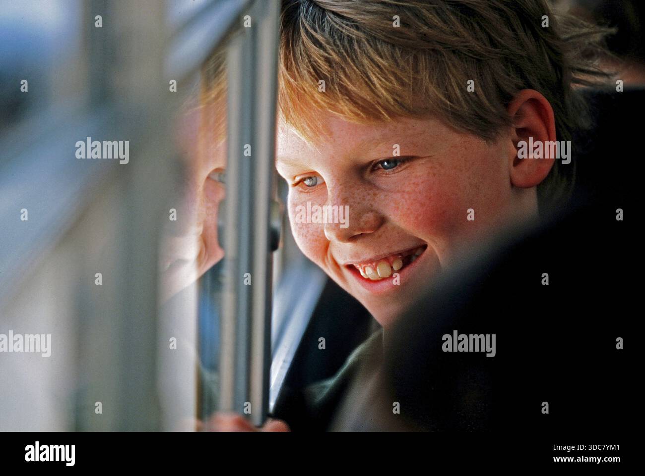 Ein Junge mit hellen Haaren lächelt, als er aus einem Busfenster schaut. Es ist ein Schulausflug, und er genießt die Aussicht und die Gesellschaft von Freunden, die in der Nähe sind Stockfoto