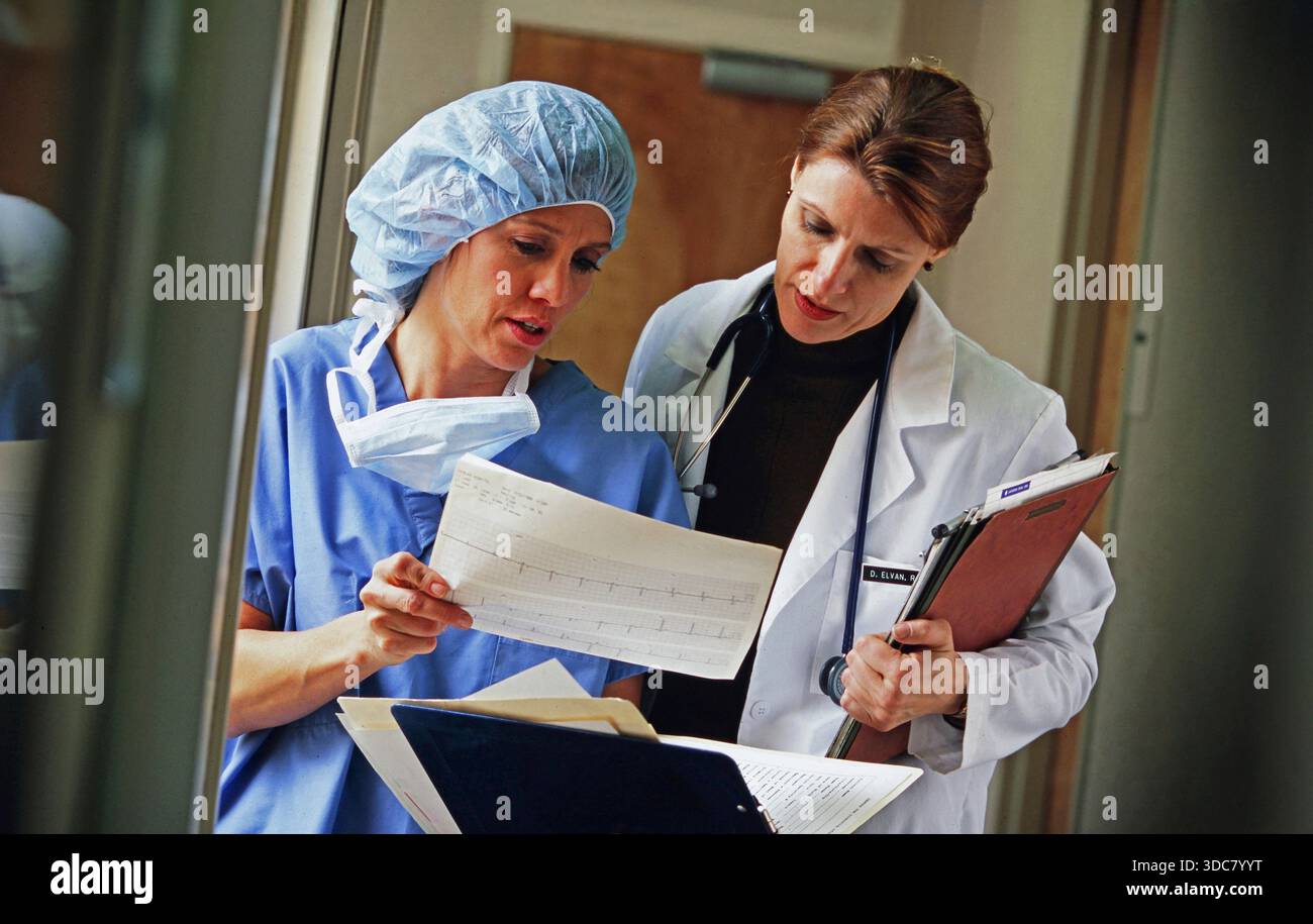 Zwei medizinische Fachkräfte diskutieren Patientenakten in einem Krankenhauskorridor. Der eine trägt Peelings und eine OP-Kappe, der andere trägt ein weißes coa Stockfoto