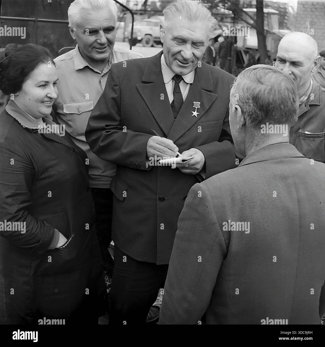 Slaviansk, Ukrainische SSR, UdSSR - 1981: Archivfoto eines Helden der Sozialistischen Arbeiterbewegung, der mit Eisenbahnarbeitern zusammentrefft. Der ältere Deputy, der einen formellen Anzug und die Goldstern-Medaille trägt, schreibt fleißig Notizen in einem Block, während er einer Frau und einem Mann in Arbeitskleidung zuhört. Die Gruppe steht in der Nähe von Bahngleisen. Dieses Bild erfasst die Interaktion zwischen der lokalen Elite und der Arbeiterklasse und unterstreicht die Reaktionsfähigkeit der Regierung in der friedlichen Donbass-Region während der späten Sowjetzeit. Stockfoto