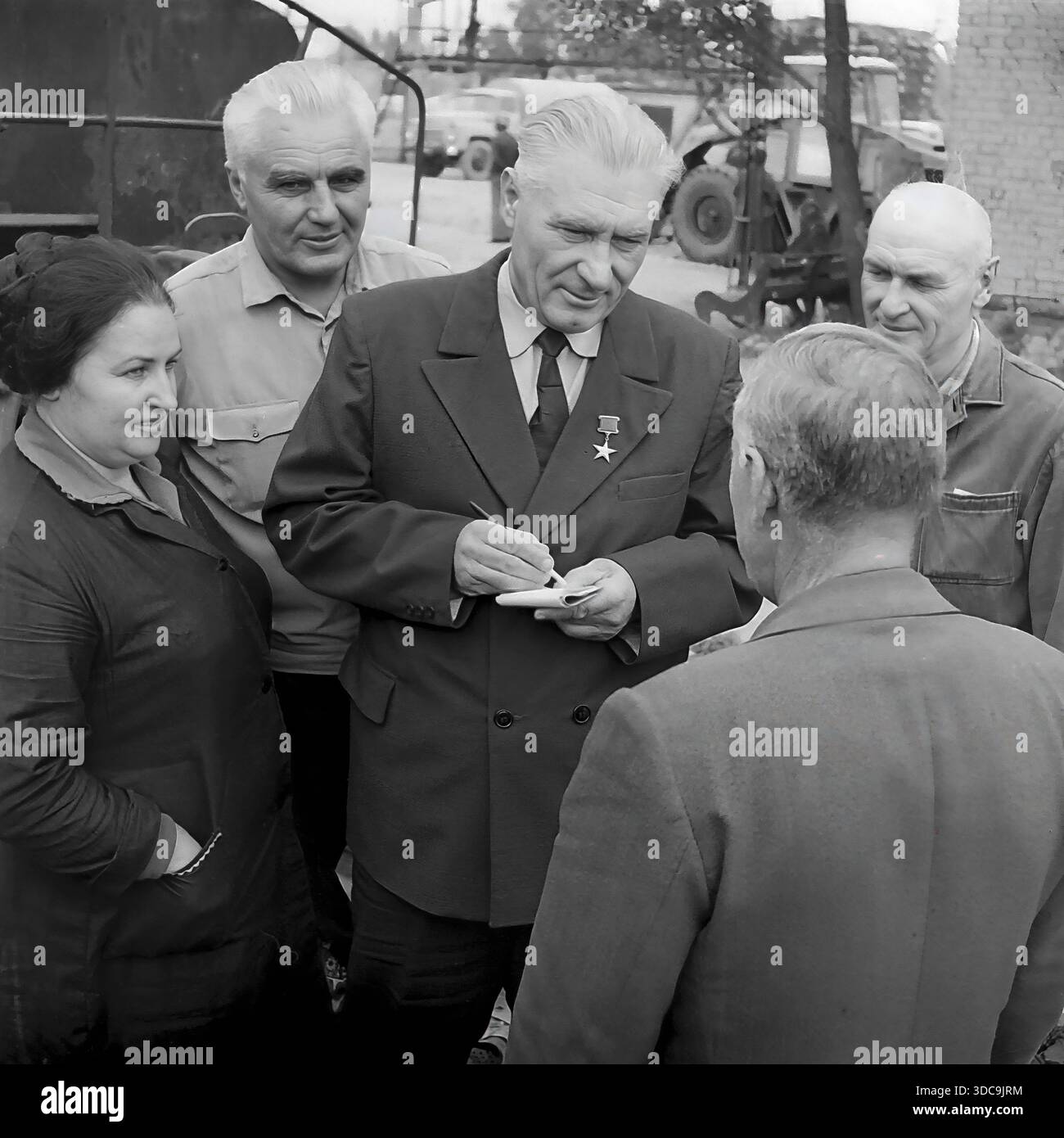 Slaviansk, Ukrainische SSR, UdSSR - 1981: Archivfoto eines Helden der Sozialistischen Arbeiterbewegung, der mit Eisenbahnarbeitern zusammentrefft. Der ältere Deputy, der einen formellen Anzug und die Goldstern-Medaille trägt, schreibt fleißig Notizen in einem Block, während er einer Frau und einem Mann in Arbeitskleidung zuhört. Die Gruppe steht in der Nähe von Bahngleisen. Dieses Bild erfasst die Interaktion zwischen der lokalen Elite und der Arbeiterklasse und unterstreicht die Reaktionsfähigkeit der Regierung in der friedlichen Donbass-Region während der späten Sowjetzeit. Stockfoto