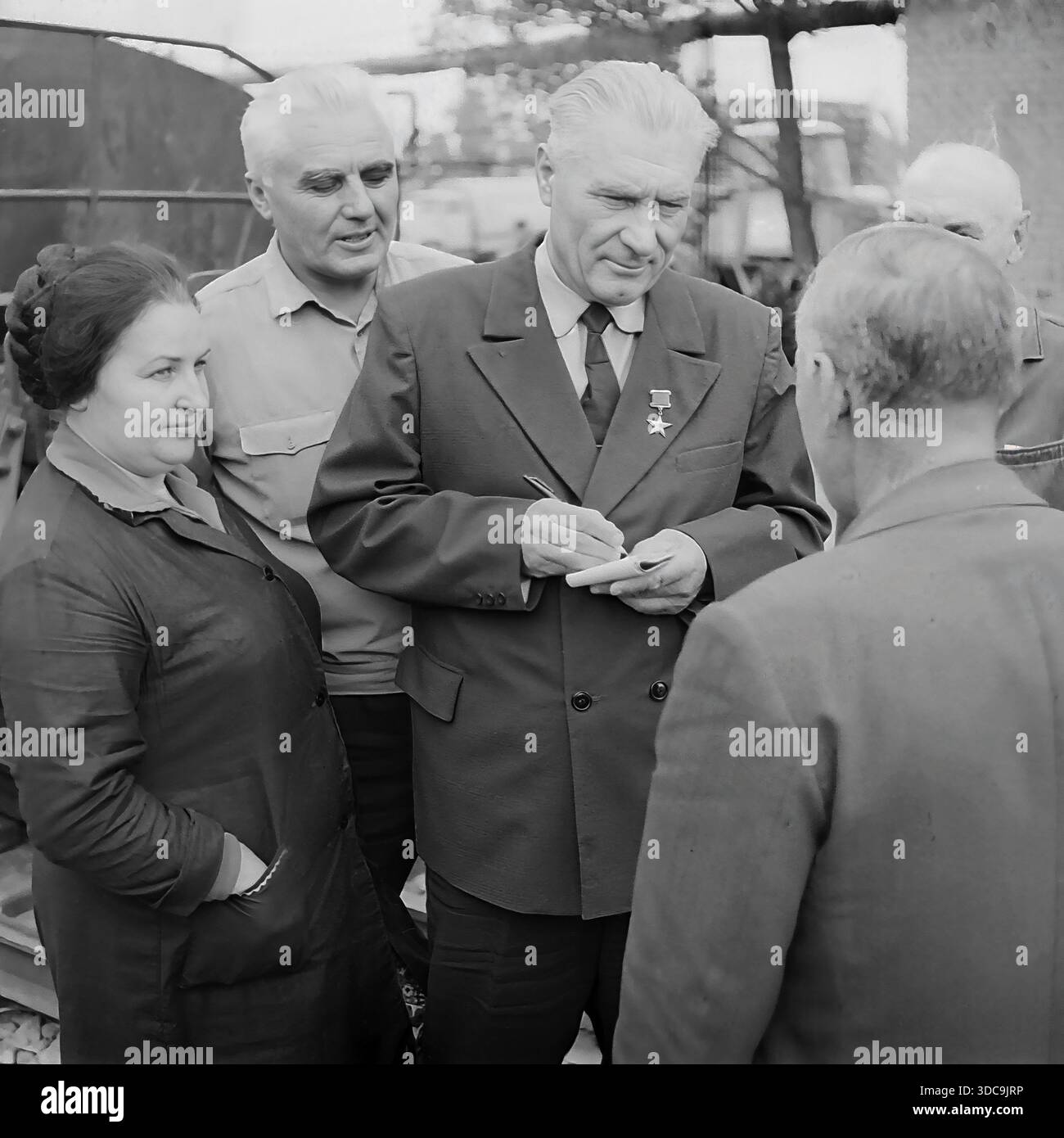 Slaviansk, Ukrainische SSR, UdSSR - 1981: Archivfoto eines Helden der Sozialistischen Arbeiterbewegung, der mit Eisenbahnarbeitern zusammentrefft. Der ältere Deputy, der einen formellen Anzug und die Goldstern-Medaille trägt, schreibt fleißig Notizen in einem Block, während er einer Frau und einem Mann in Arbeitskleidung zuhört. Die Gruppe steht in der Nähe von Bahngleisen. Dieses Bild erfasst die Interaktion zwischen der lokalen Elite und der Arbeiterklasse und unterstreicht die Reaktionsfähigkeit der Regierung in der friedlichen Donbass-Region während der späten Sowjetzeit. Stockfoto