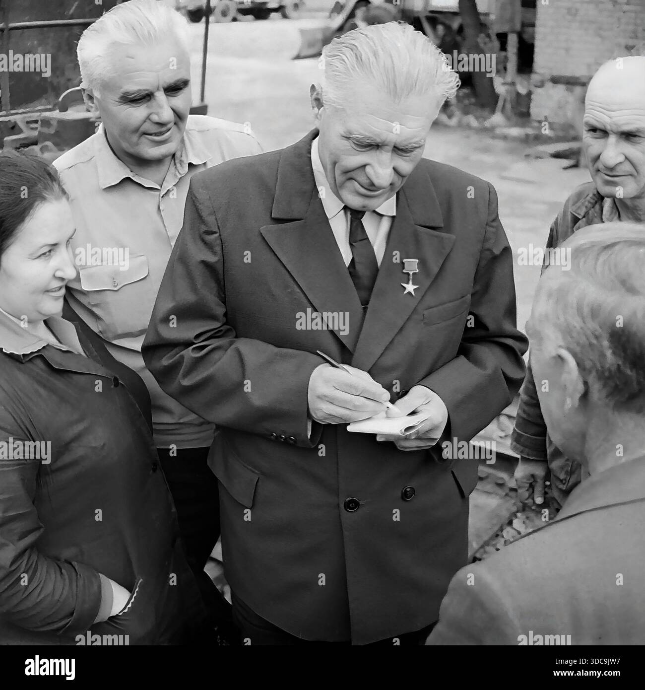 Slaviansk, Ukrainische SSR, UdSSR - 1981: Archivfoto eines Helden der Sozialistischen Arbeiterbewegung, der mit Eisenbahnarbeitern zusammentrefft. Der ältere Deputy, der einen formellen Anzug und die Goldstern-Medaille trägt, schreibt fleißig Notizen in einem Block, während er einer Frau und einem Mann in Arbeitskleidung zuhört. Die Gruppe steht in der Nähe von Bahngleisen. Dieses Bild erfasst die Interaktion zwischen der lokalen Elite und der Arbeiterklasse und unterstreicht die Reaktionsfähigkeit der Regierung in der friedlichen Donbass-Region während der späten Sowjetzeit. Stockfoto