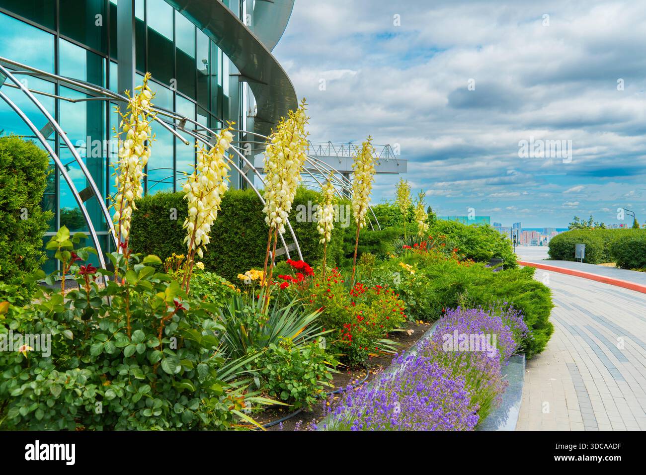 Malerischer Blick auf eine moderne Glasarchitektur-Fassade mit einem lebhaften Garten mit blühenden Yucca, Rosen und Lavendel unter bewölktem Himmel in einem Stadtpark Stockfoto