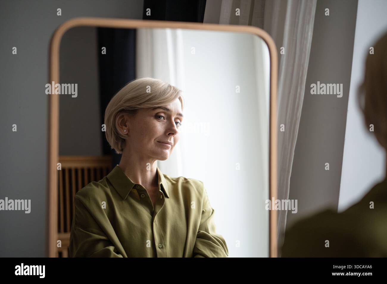 Schöne blonde Frau mittleren Alters, die vor dem Spiegel steht und lächelt und sich selbst reflektiert. Stockfoto