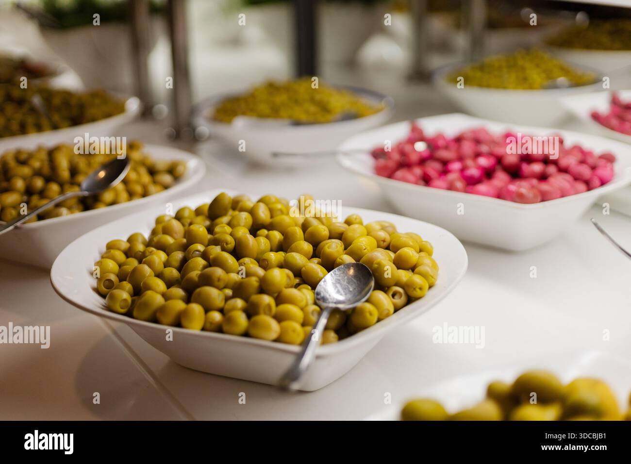 Eine Auswahl an Oliven wird in Schüsseln am Buffet serviert. Es werden grüne und rosa Oliven ausgestellt. Einige Schüsseln haben Servierlöffel in der Nähe für g Stockfoto