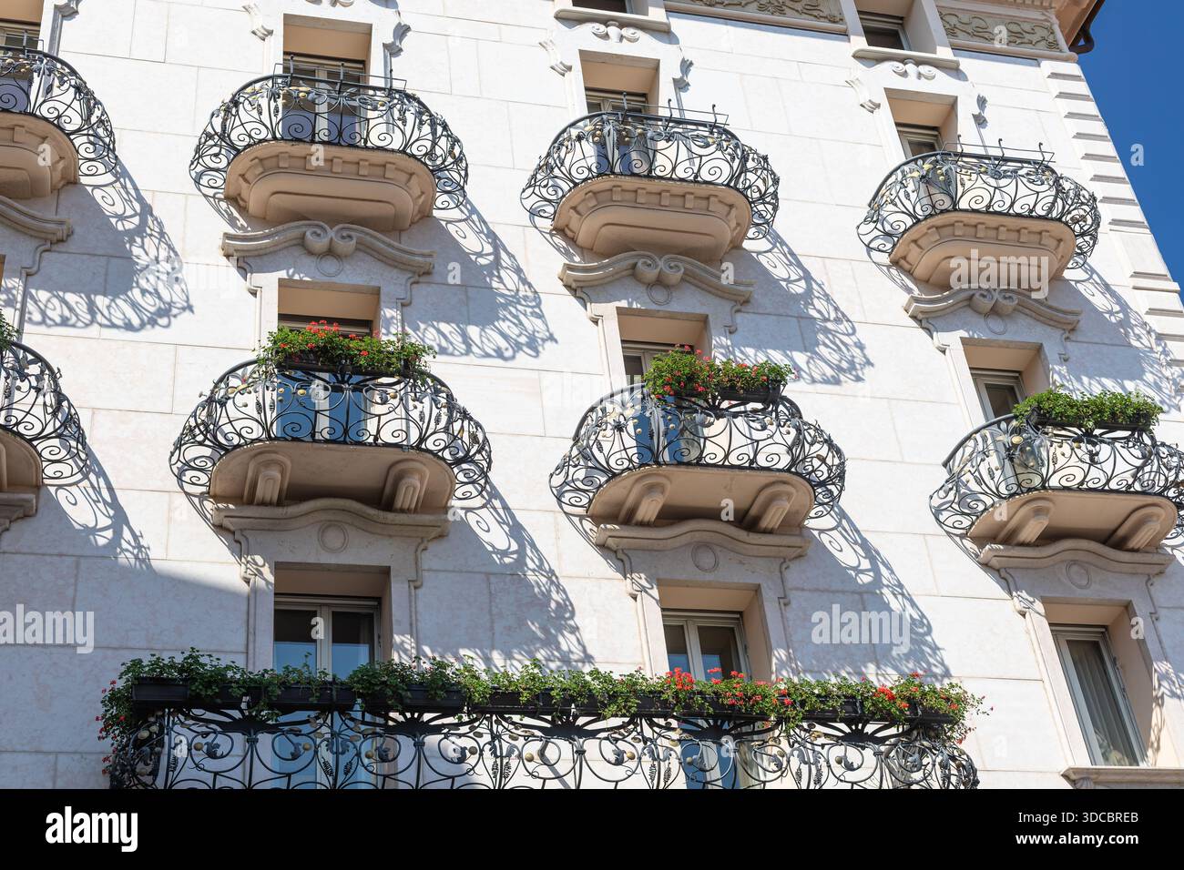 Die sonnendurchflutete weiße Fassade verfügt über dekorative Eisenbalkone mit Blumenkästen, die architektonische Details, Schattenmuster und urbanen Charme zeigen. Stockfoto