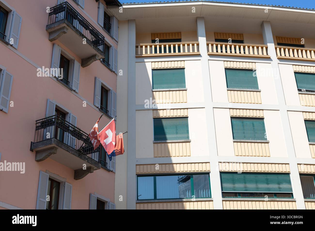 Eine helle Stadtfassade zeigt eine Schweizer Flagge, die von einem Balkon eines rosafarbenen Gebäudes mit grünen Fensterläden hängt, die europäische Architektur einfängt, in Farbtönen gehalten Stockfoto