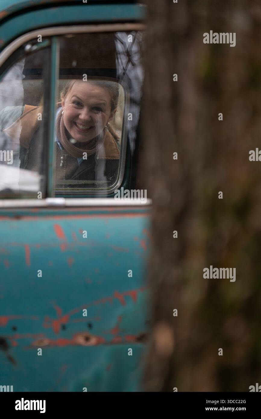 Lächelnde Frau schaut durch das Fenster eines Oldtimer-türkisfarbenen Autos, abblätternde Farbe, sanftes Herbstlicht, flache Tiefen des Feldes, warme nostalgische Stimmung Stockfoto