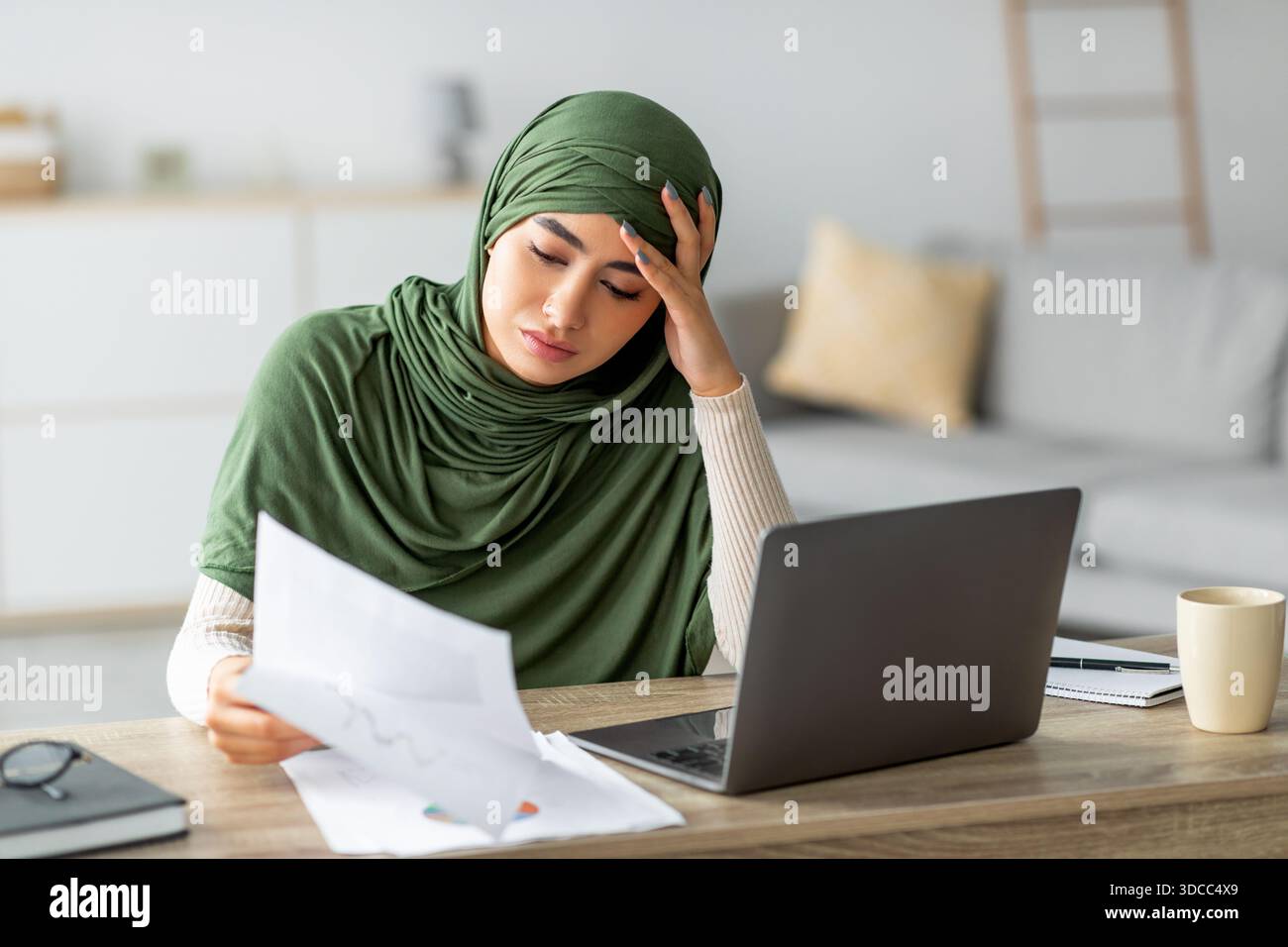 Eine konzentrierte junge arabische Frau liest Dokumente, während sie an einem Online-Geschäftstreffen in ihrem Heimbüro teilnimmt. Sie strahlt Entschlossenheit aus, während sie navigiert Stockfoto