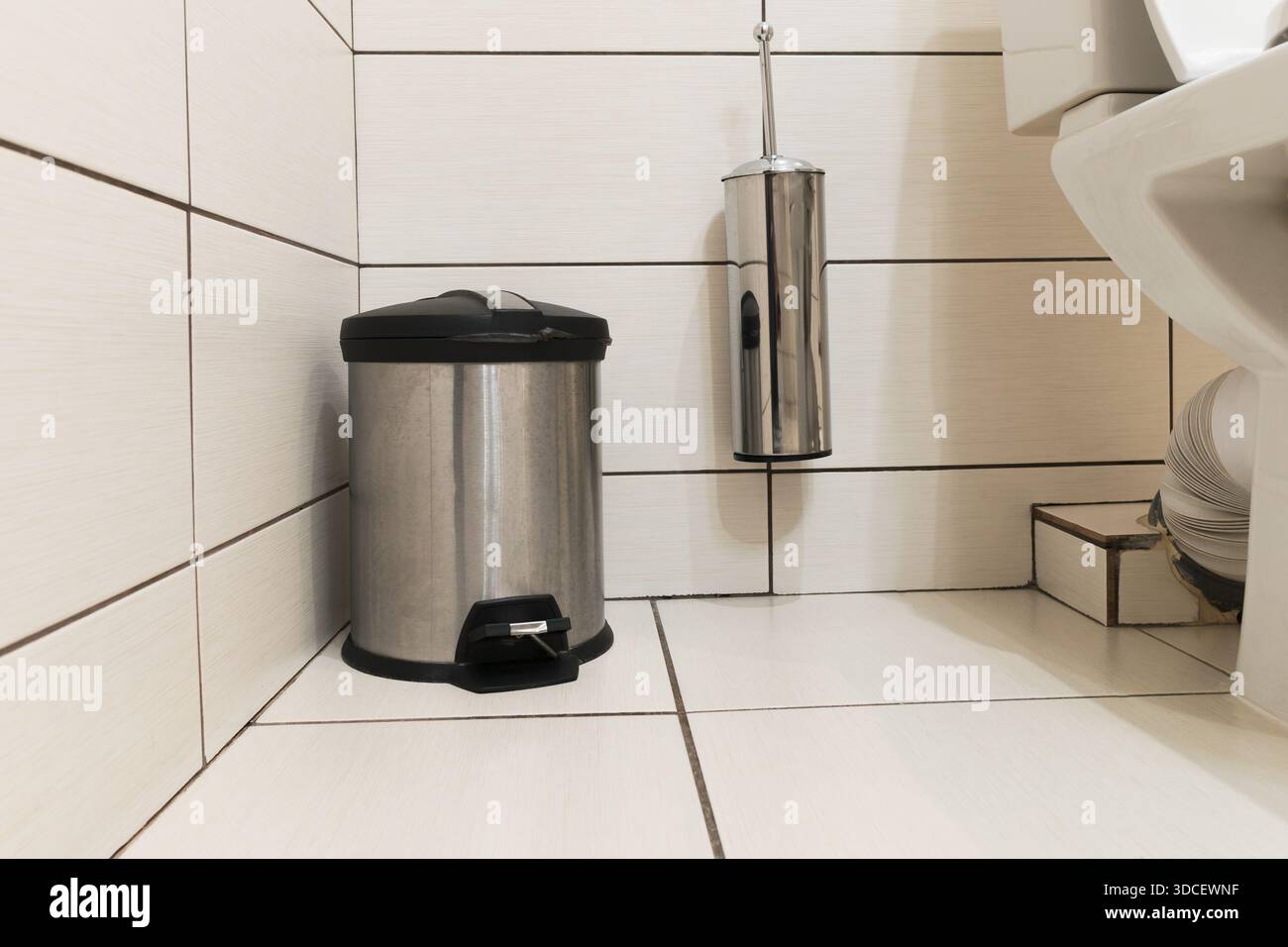 Mülleimer aus Edelstahl mit Fußpedal auf dem Boden und einer wandmontierten Toilettenbürste in einem Badezimmer mit großen hellen Keramikfliesen Stockfoto