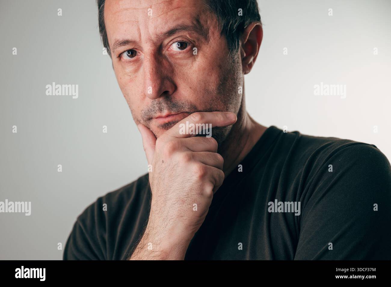 Porträt eines Mannes mittleren Alters mit kurzen dunklen Haaren und Stoppeln, der nachdenklich mit Hand am Kinn vor neutralgrauem Hintergrund blickt. Reflexion, wor Stockfoto