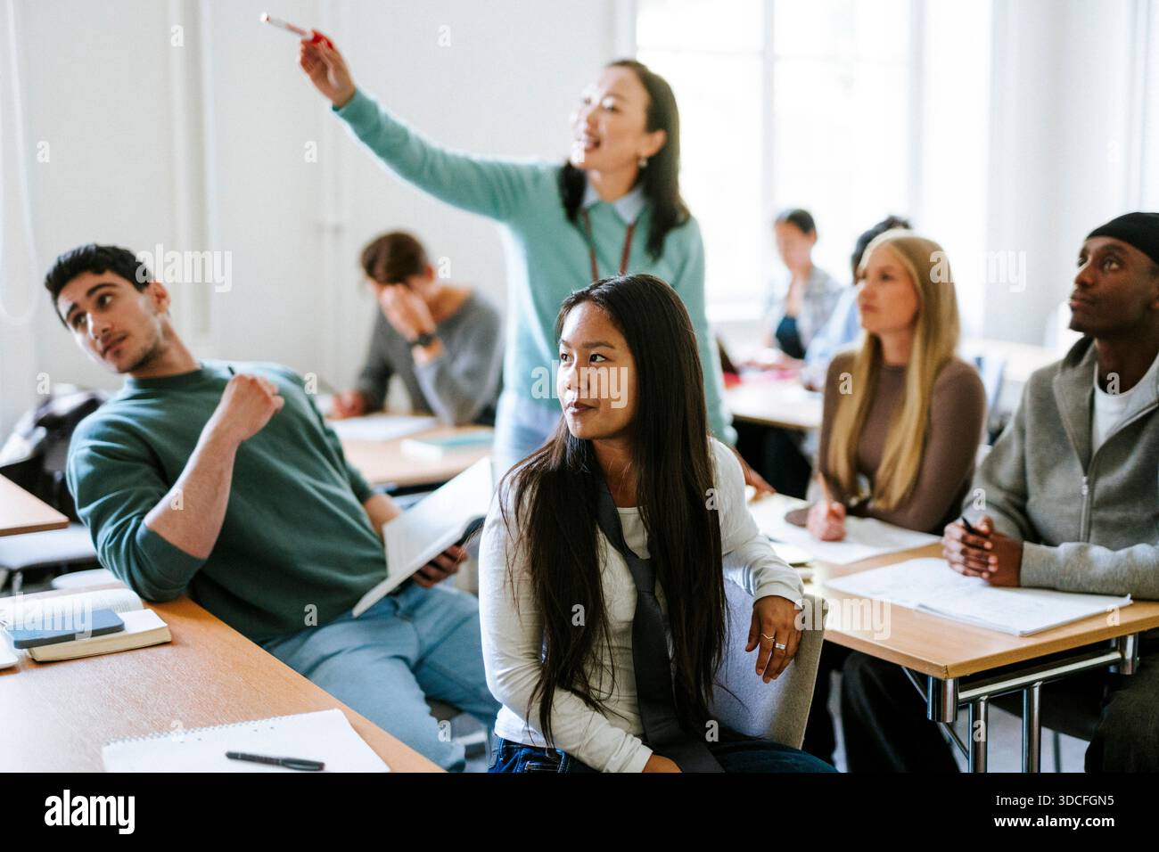 Langhaarige junge Frau, die dem Lehrer zuhört, während sie am Schreibtisch im Klassenzimmer am College sitzt Stockfoto