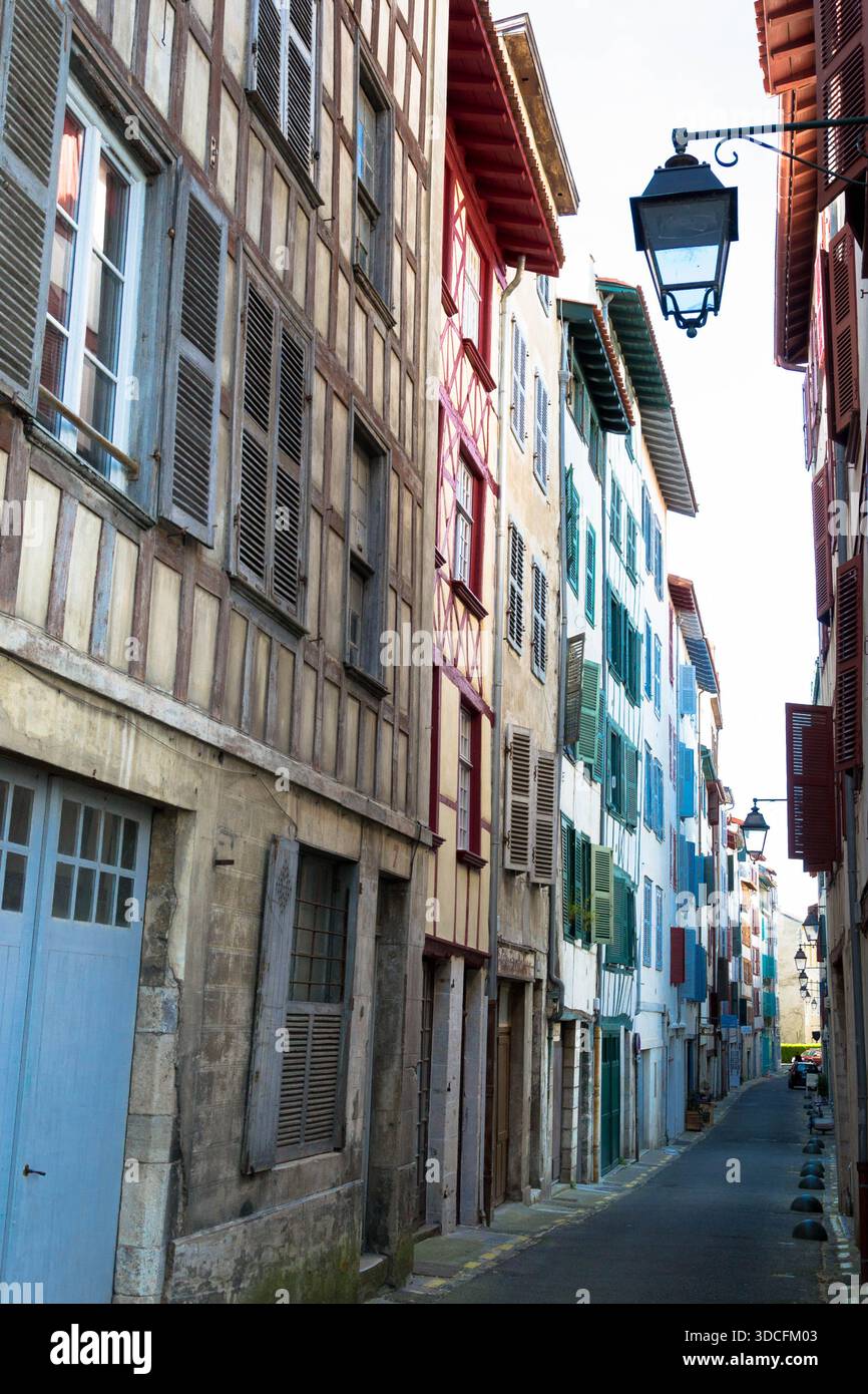 Blick auf farbenfrohe Gebäude in einer engen Straße, mit lebhaften Fensterläden und malerischen Straßenlaternen, die Schatten auf den Bürgersteig werfen, Bayonne, F Stockfoto
