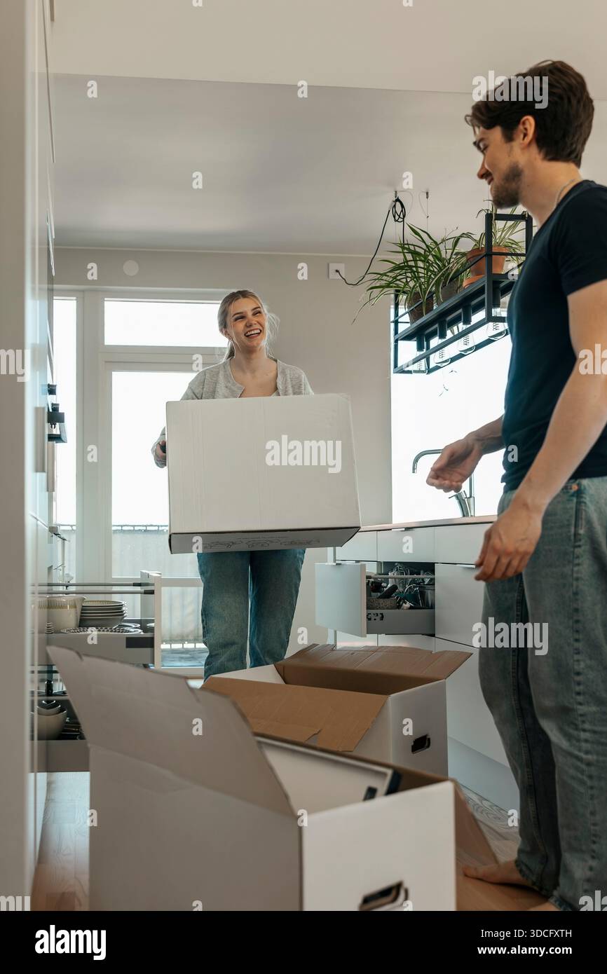 Lächelnde junge Frau, die Pappkarton beim Auspacken von Sachen mit Freund im neuen Haus trägt Stockfoto